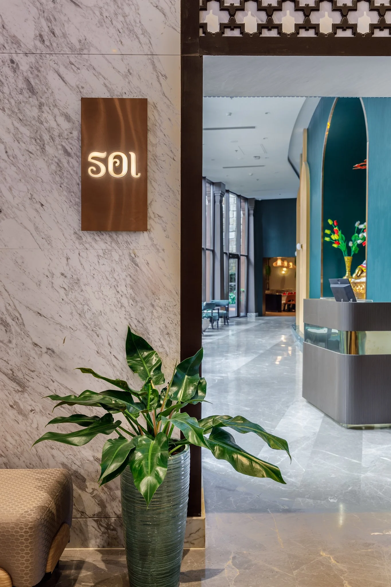 Interior view of a hotel lobby with a large green potted plant, marble walls, a reception desk, and a digital sign displaying the number 502.