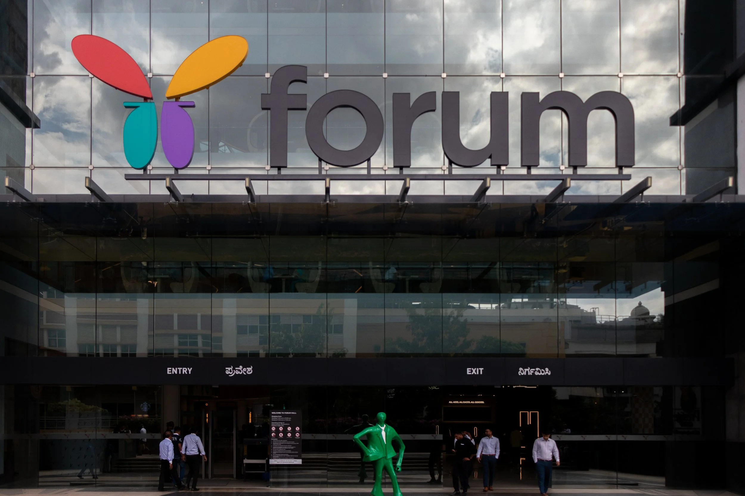 Front view of a modern building with large glass windows and a sign that reads 'forum' in lowercase letters. The logo above the sign has four colorful shapes in red, yellow, teal, and purple. Several people are walking in front of the entrance, and t