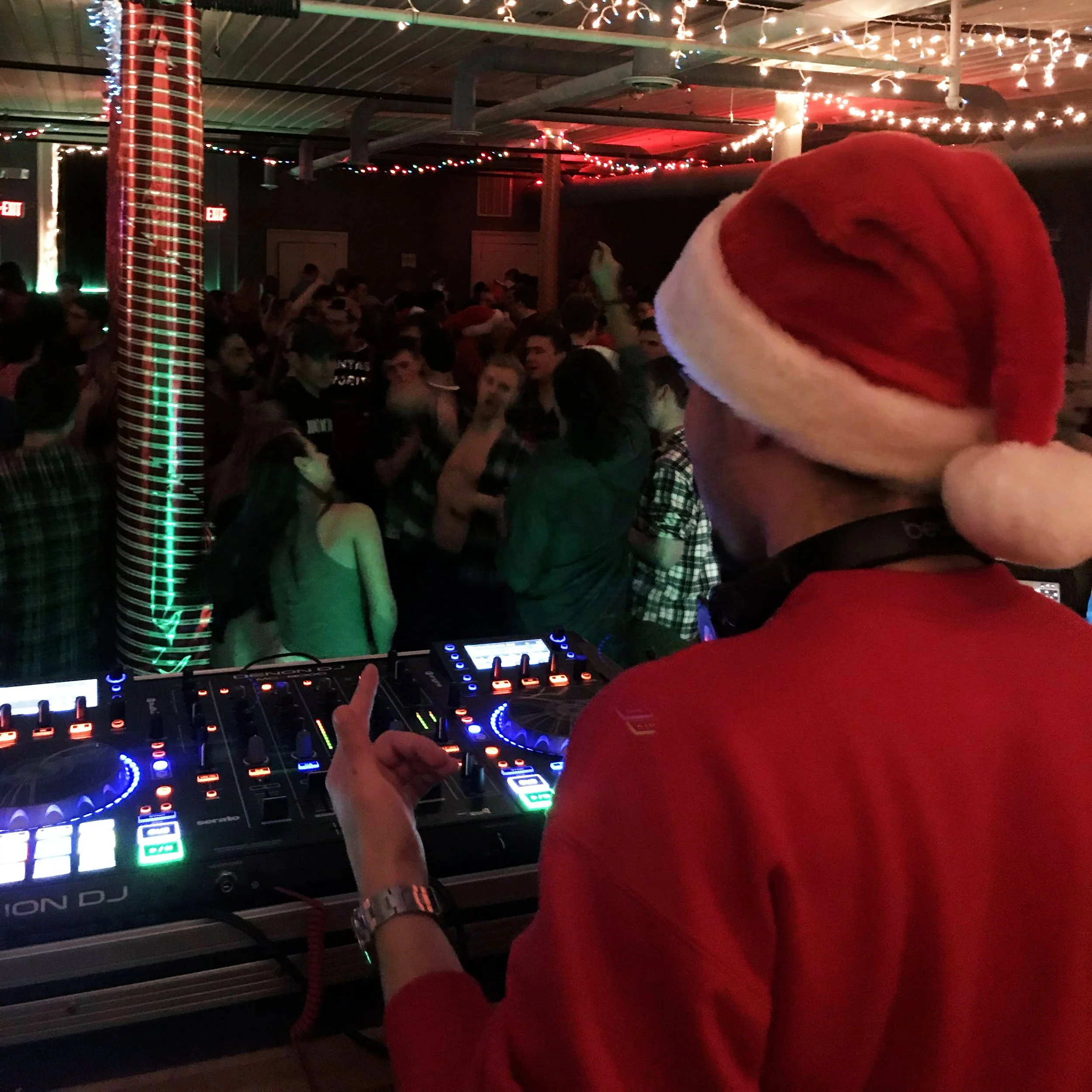 An image from behind Mykroh as he DJs to a lively dark room of dancing people for a Christmas party. In the foreground is a Denon MCX8000, as well as pillars wrapped in gift wrap and string lights.