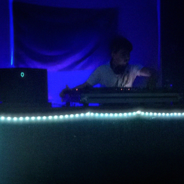 An image of a young mykroh DJing in a white tee shirt next to an Alienware Laptop with string lights and a blank flag behind him.