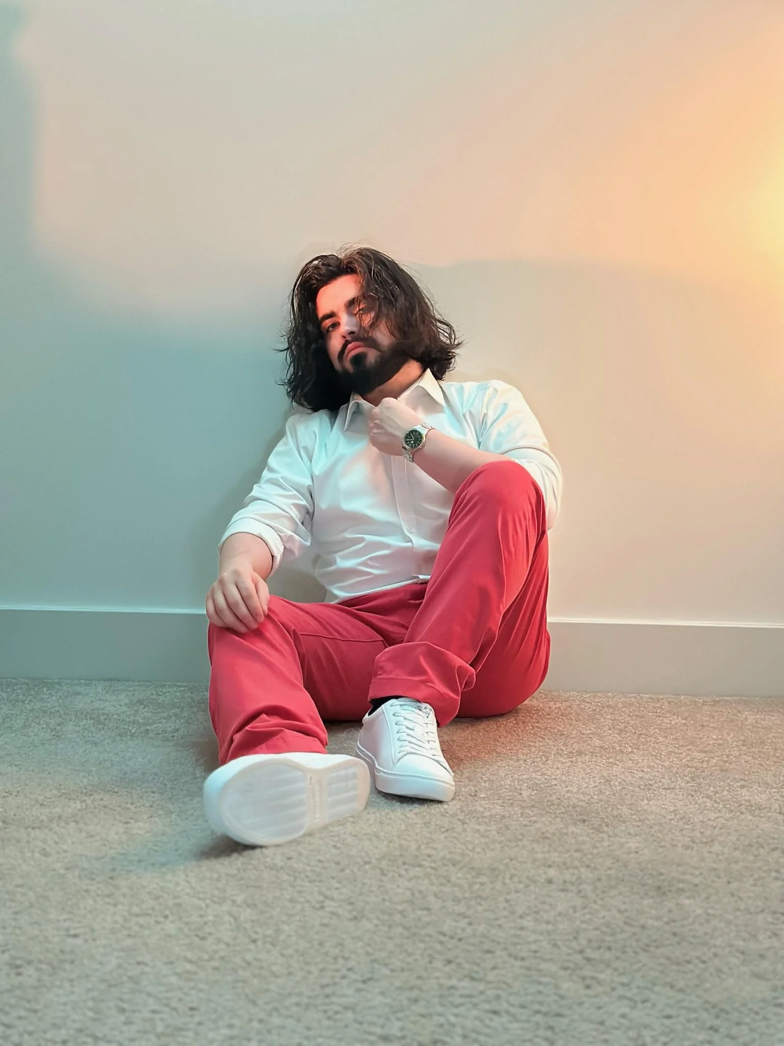 a photoshoot image of Mykroh in semi-formal clothing and white sneakers, seated against a wall clasping his collar with his left arm