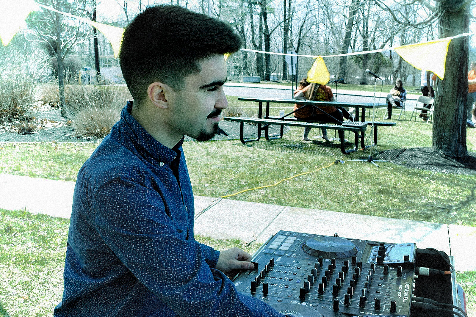 An image of Mykroh DJing a block party for charity with yellow flags on a white string behind him, with a Denon MCX8000 DJ controller in front of him.