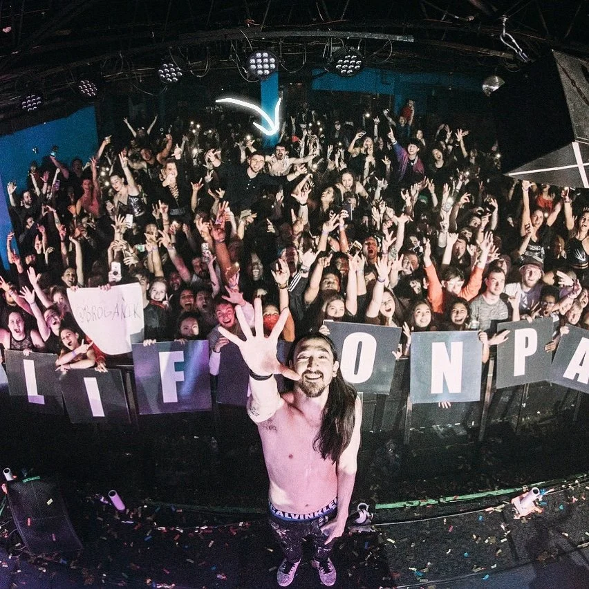 An image of Steve Aoki in front of a crowd with a young Mykroh crowd surfing with a white glowing arrow pointing to him.