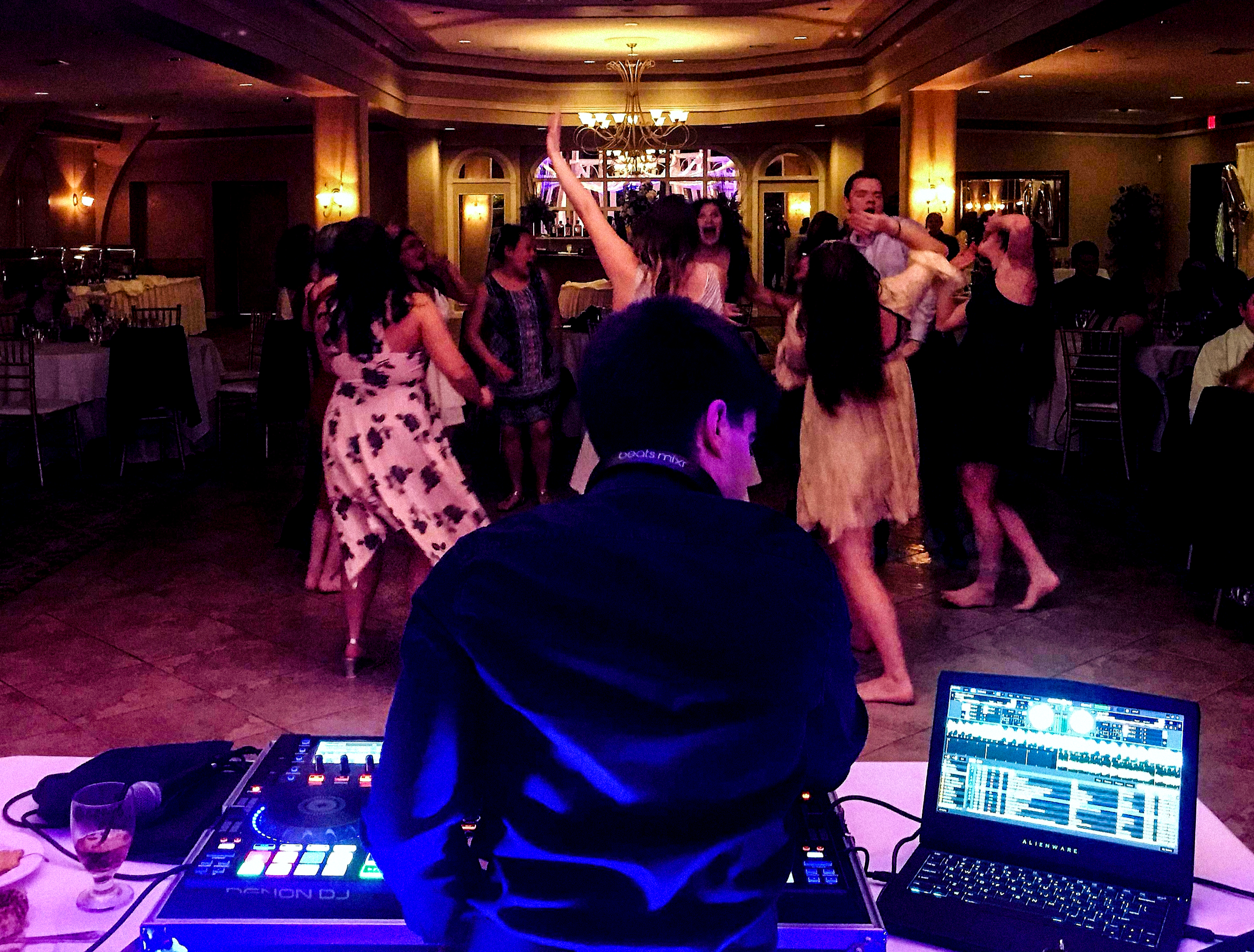 An image of Mykroh DJing to a lively dance floor with mostly young women in semi-formal dress lit by reddish-pink lighting.