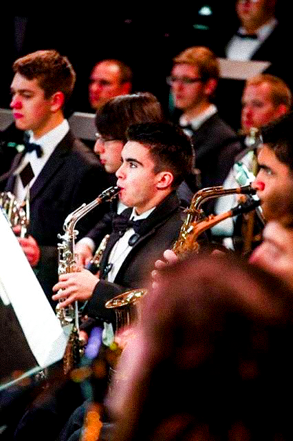 An image of Mykroh in a black suit, playing a silver alto saxophone with other instrumentalists around him