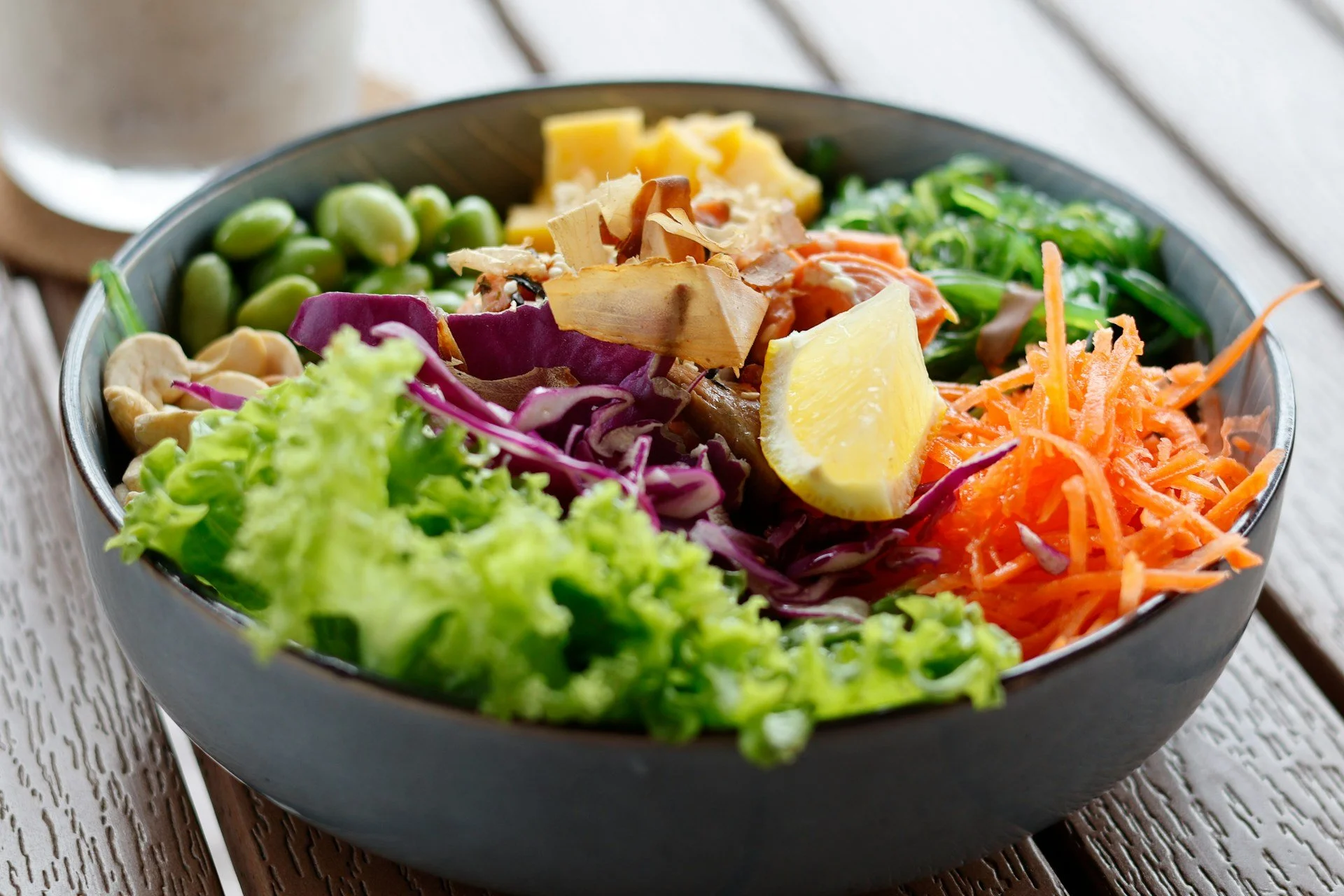 A close-up of a bowl of fresh green leaves, sliced red cabbage, nuts, edamame, seaweeds, shredded carrots and a cut of lemon, arranged in a vibrant and colourful presentation, perfect for Spring dish.