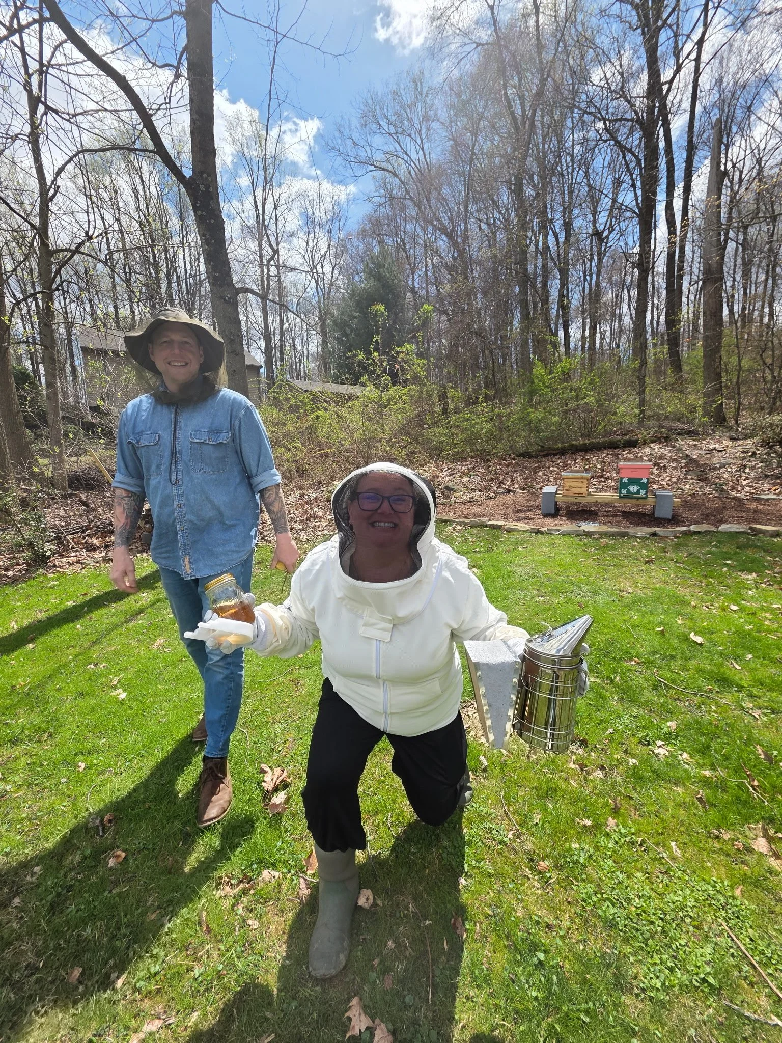 Two women and a woman. The woman at front is kneeling, wearing a white jacket, gloves, and rain boots, holding a jar of honey. The woman at back is standing, wearing a hat and denim jacket. They are outdoors in a wooded area with blue sky and trees in the background.