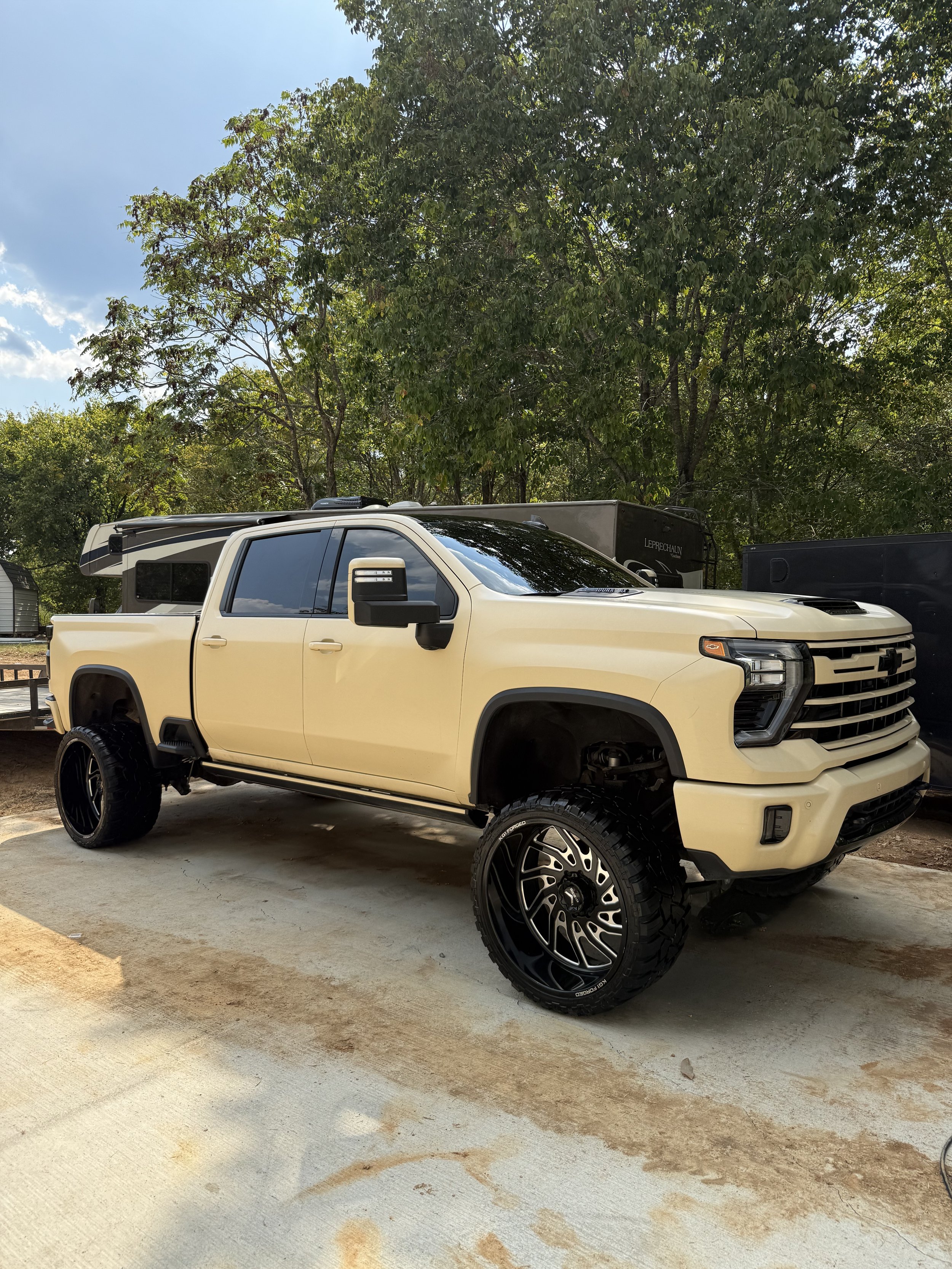 A beige lifted pickup truck with large off-road tires parked outdoors under a blue sky with trees in the background.