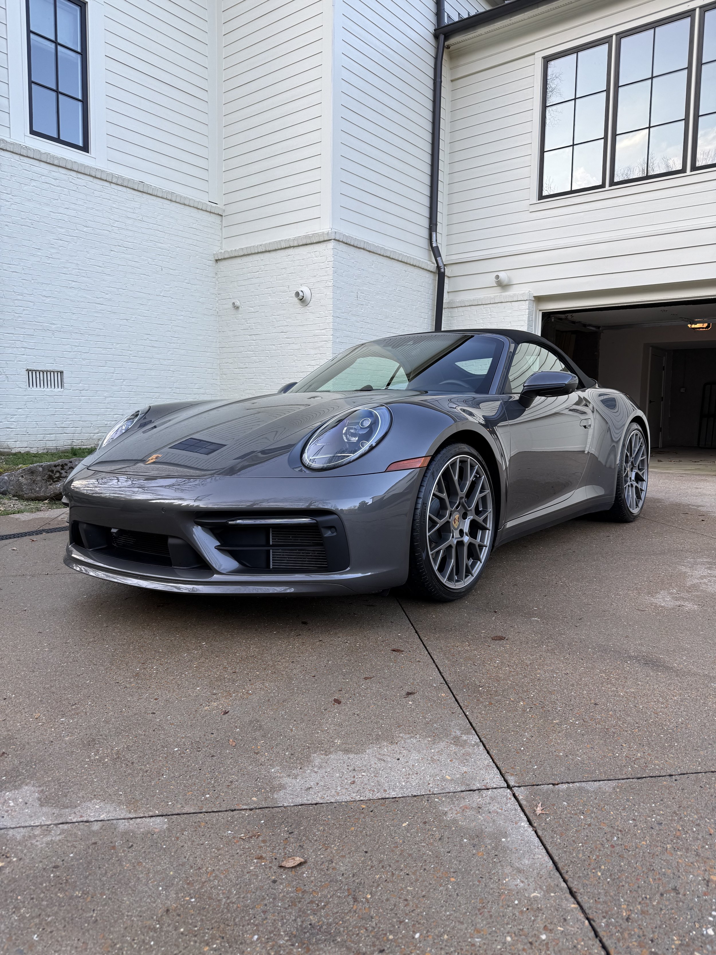 Gray Porsche 911 convertible parked on a driveway in front of a white house with large windows and a garage.