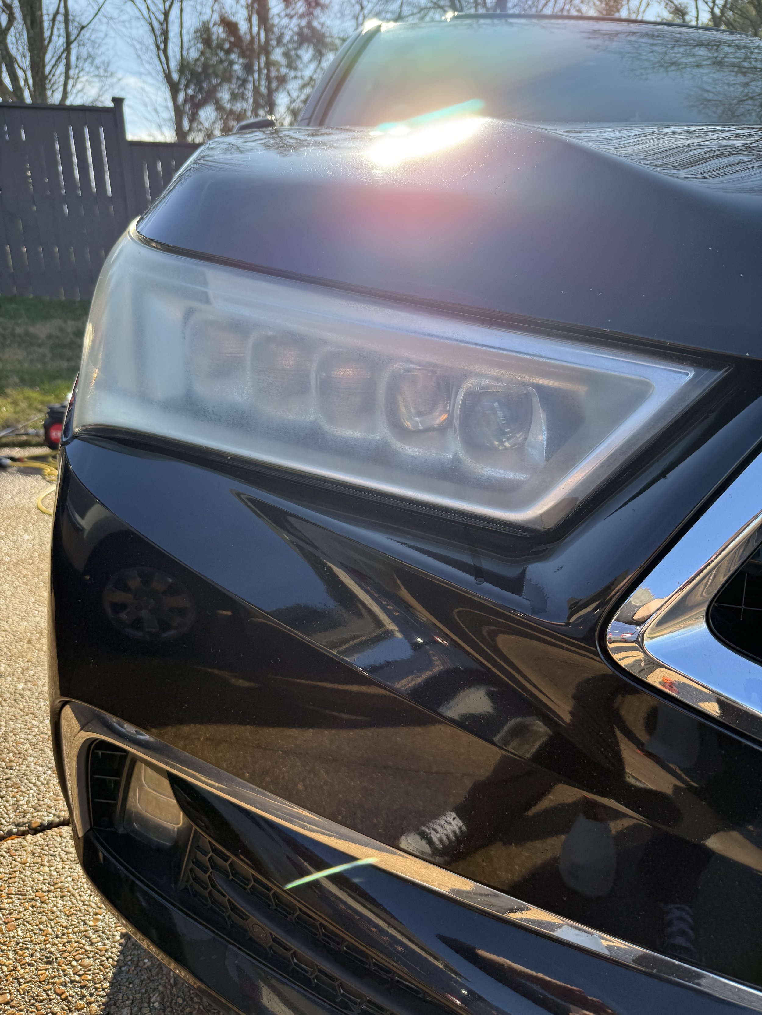 Close-up of the front right headlight and bumper of a black car parked on a driveway, with sunlight glare and reflections visible on the car surface.