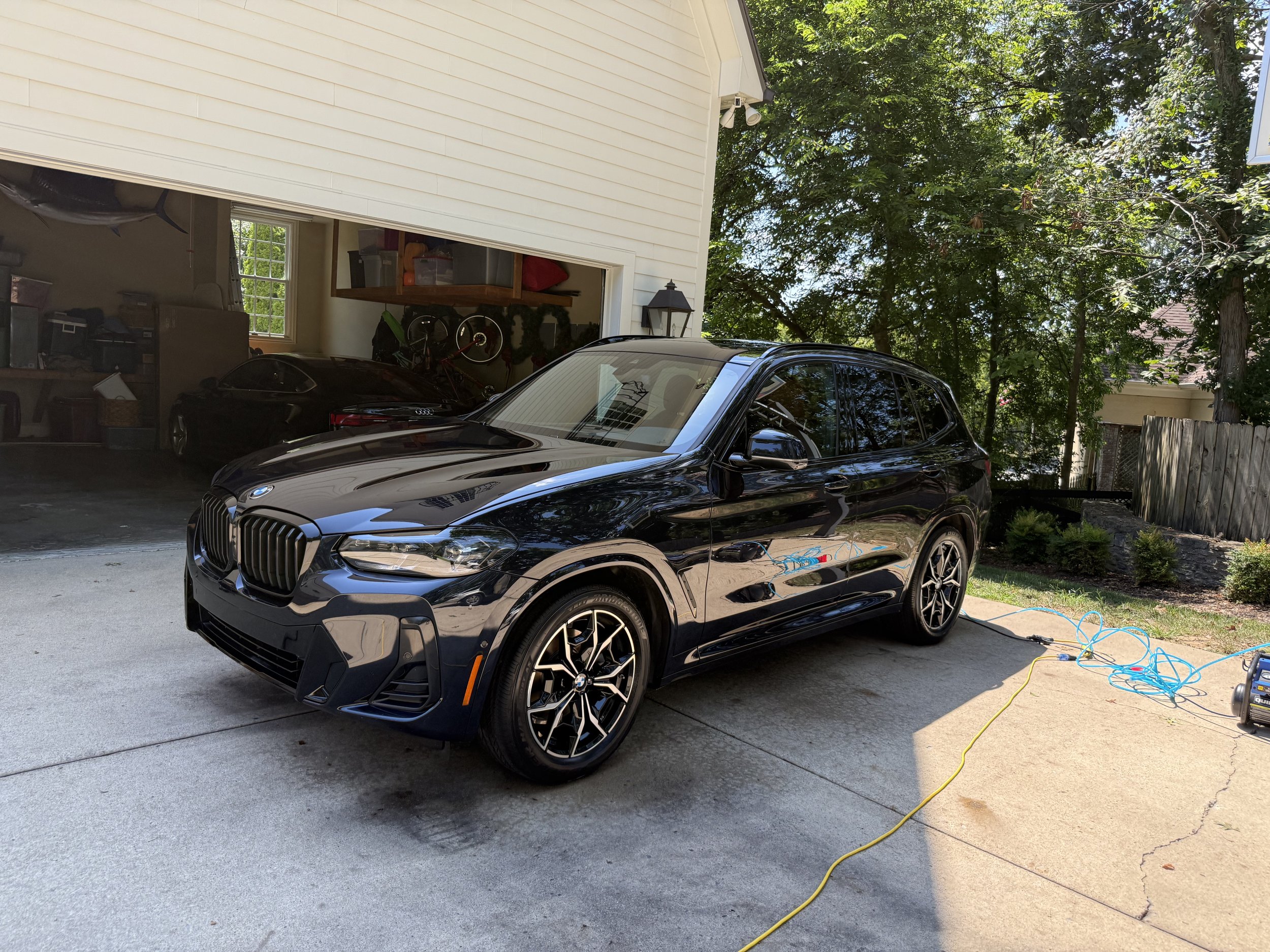 Black BMW SUV parked in driveway outside garage with a yellow and blue power cord connected to an electric device on the ground. Inside the garage, a black car is partially visible along with bicycles, storage boxes, and shelving.