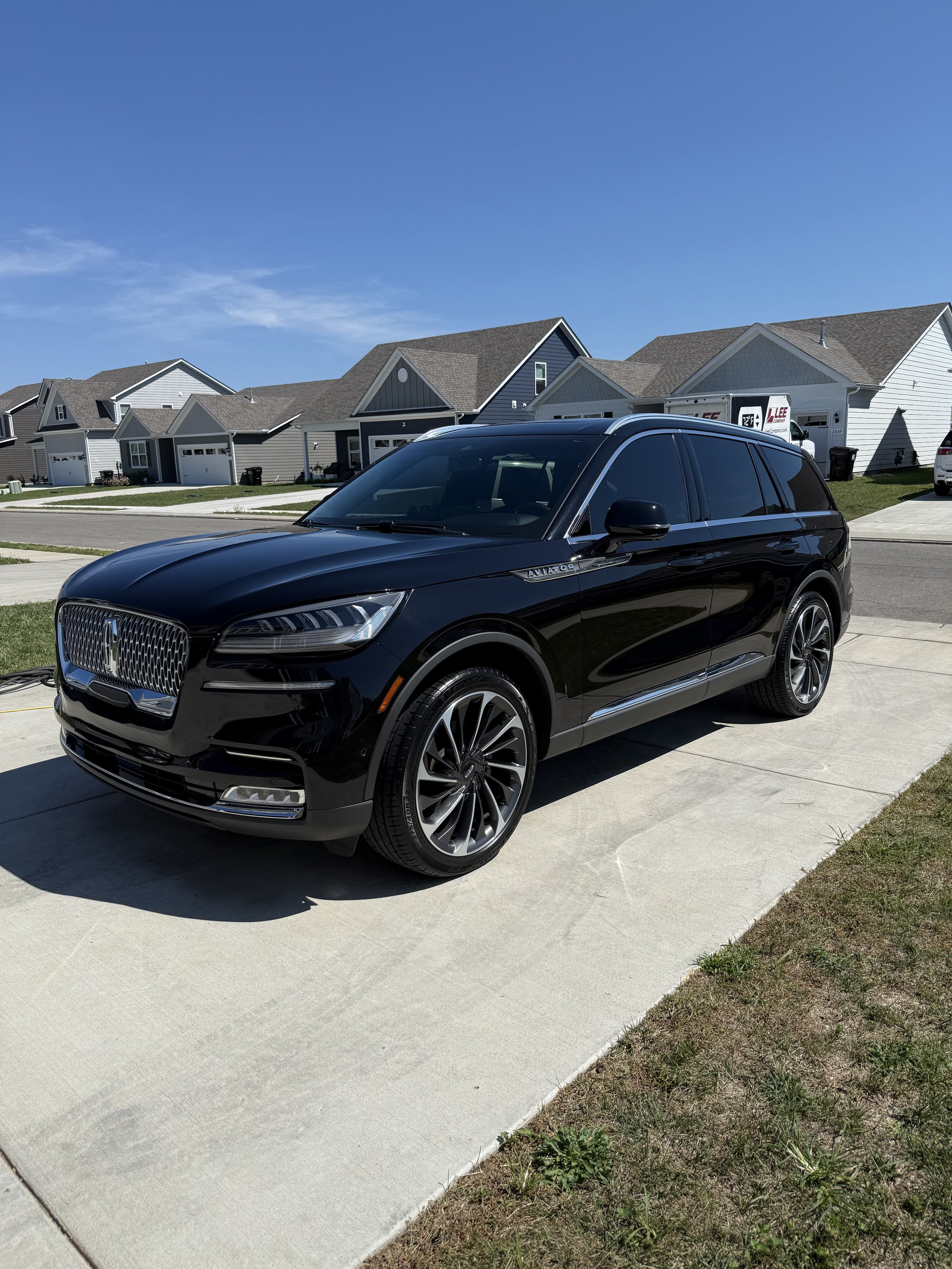 A black Lincoln Aviator SUV parked on a driveway in a suburban neighborhood with row of houses under a clear blue sky.