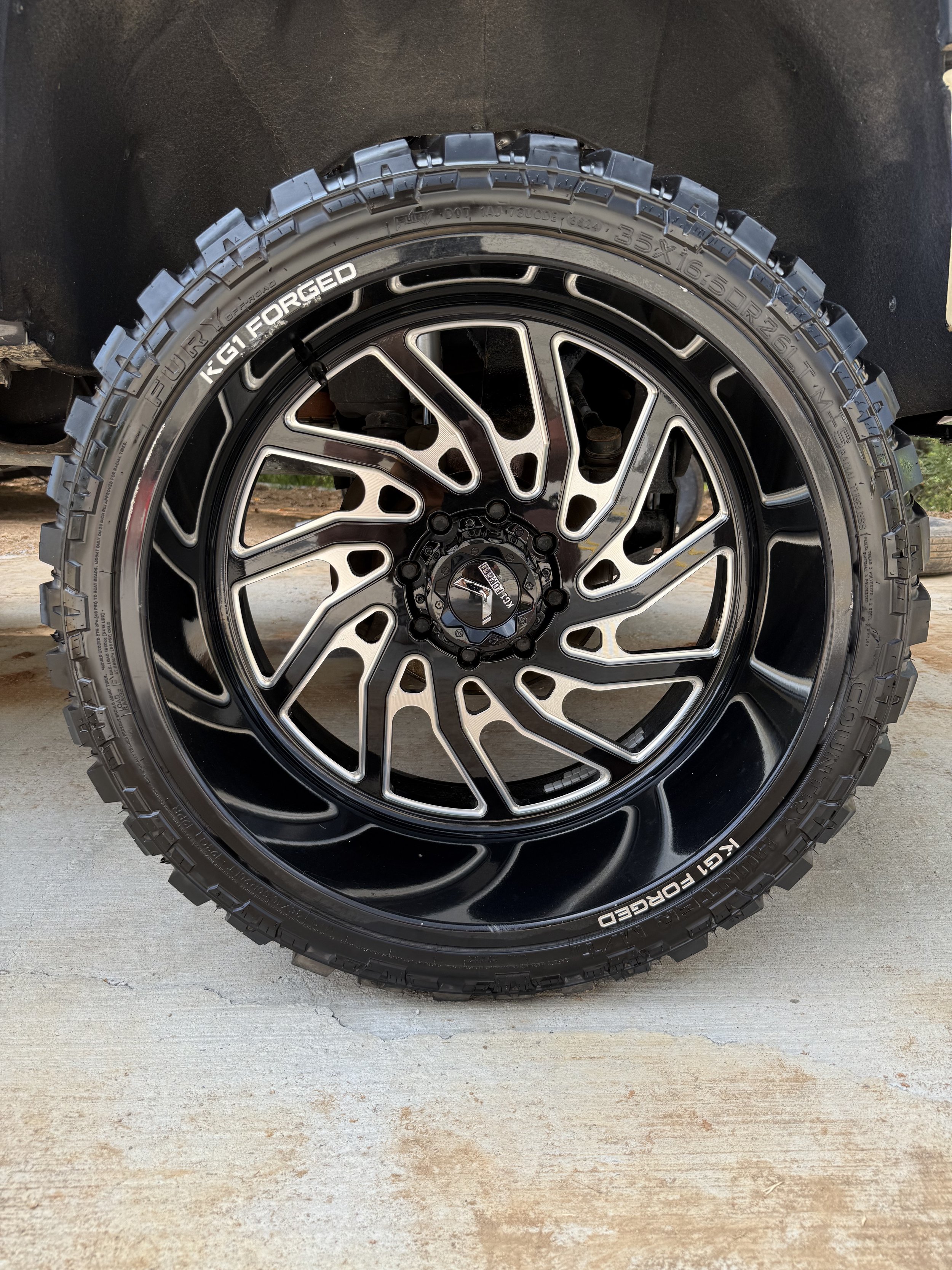 Close-up of a black off-road tire with aggressive tread, mounted on a black and silver alloy wheel, on a concrete surface.
