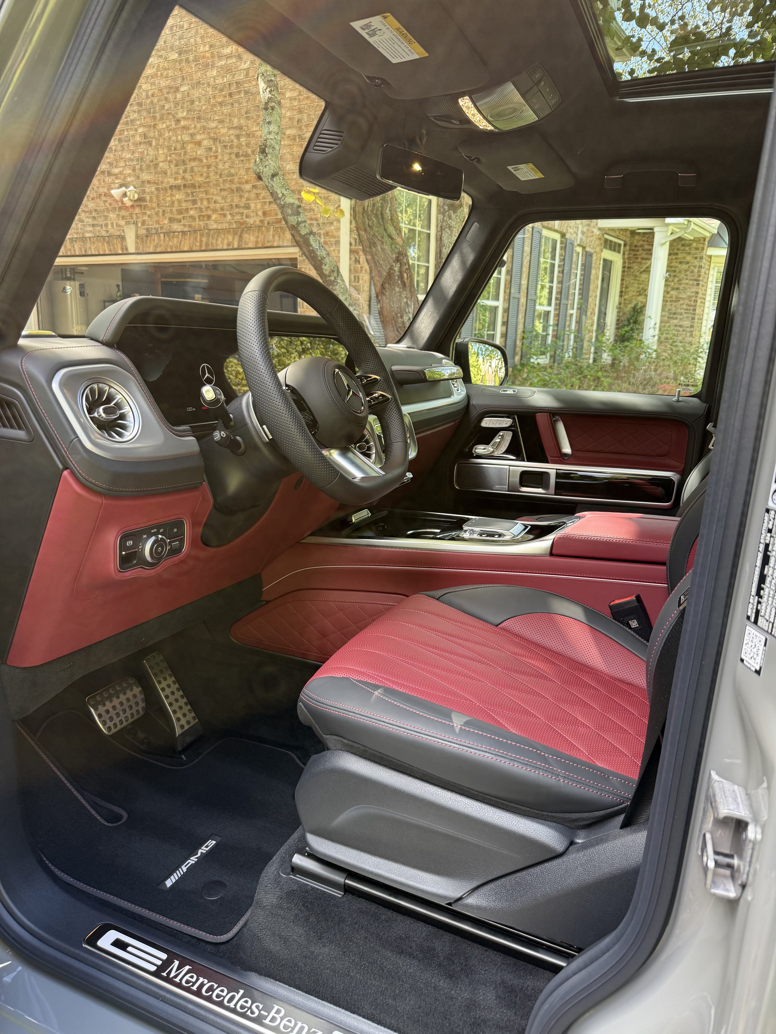 Interior view of a Mercedes-Benz G-Class, featuring a red and black leather seat, black dashboard with Mercedes logo on steering wheel, and a sunroof, with a brick house visible through the window.