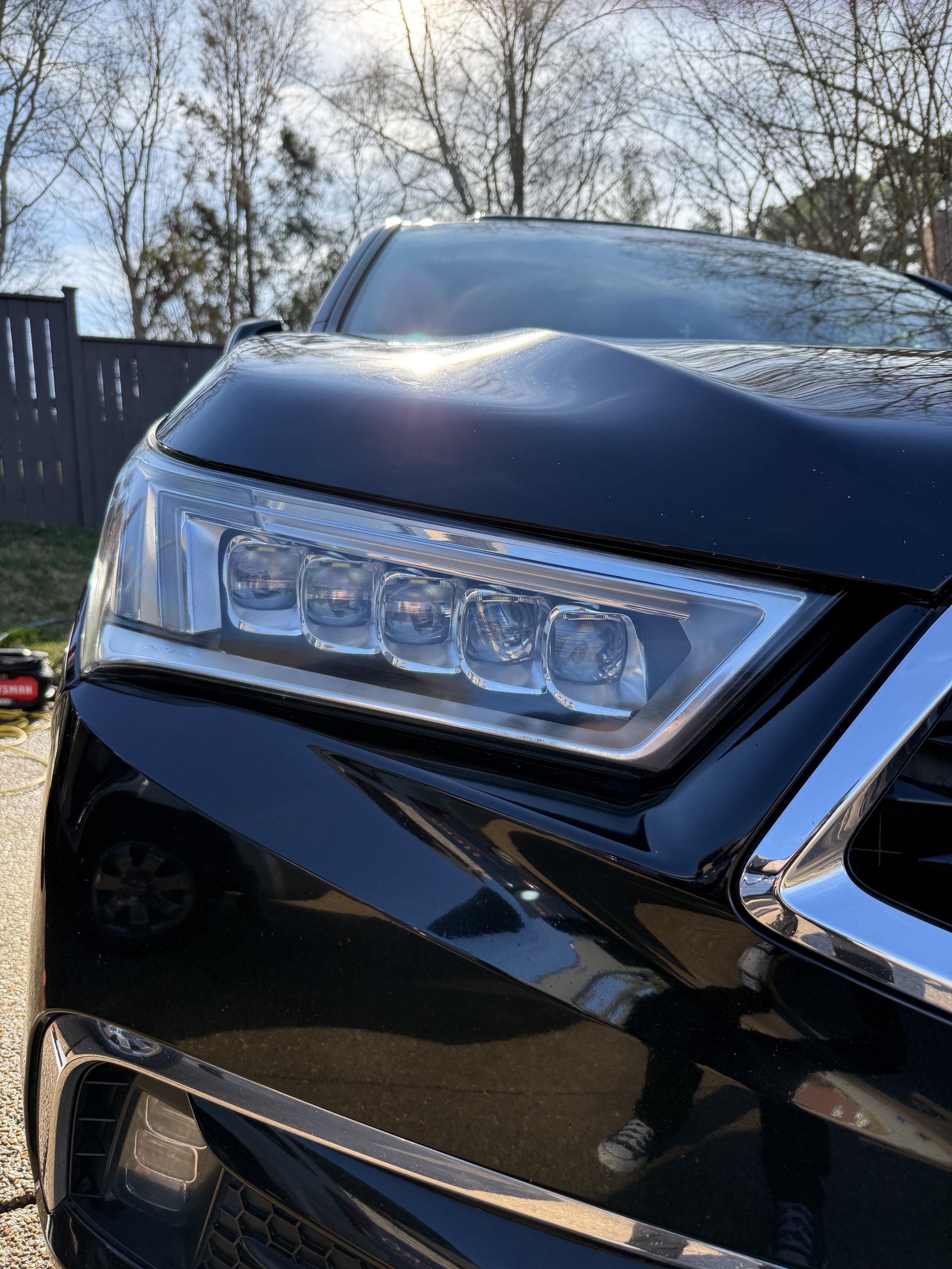 Close-up of the front end of a black modern car with LED headlights, parked outdoors with clear sky and leafless trees in the background.
