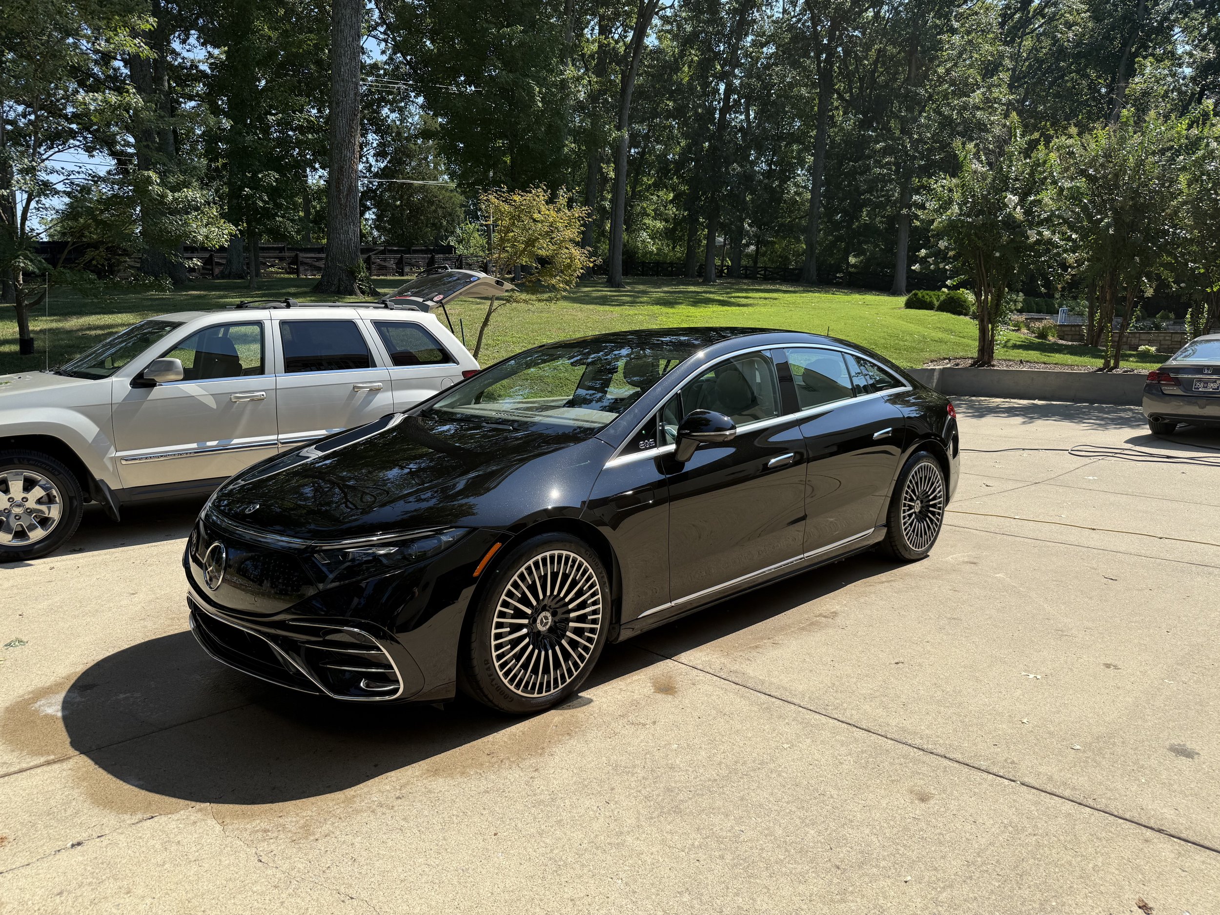 Black Mercedes-Benz vehicle parked on a driveway during daytime with other cars and green trees in the background.