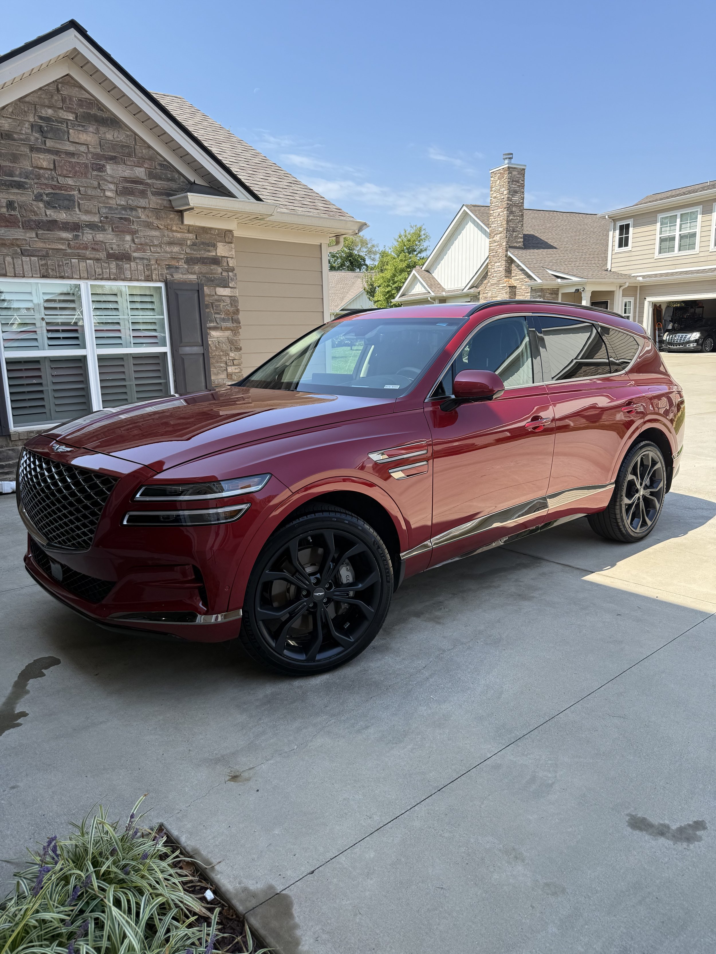 Red SUV parked on a concrete driveway in front of a house with a brick and siding exterior, blue sky, and neighboring houses visible in the background.