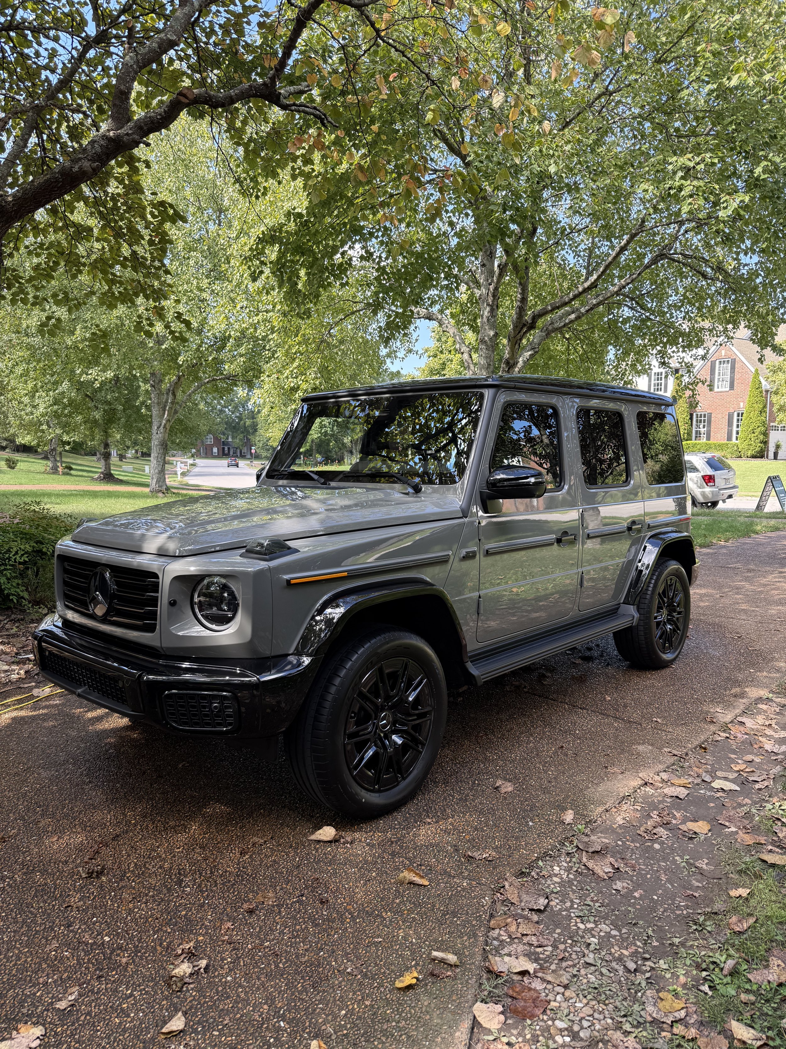 A silver Mercedes-Benz G-Class SUV parked on a gravel path under green trees with a neighborhood in the background.