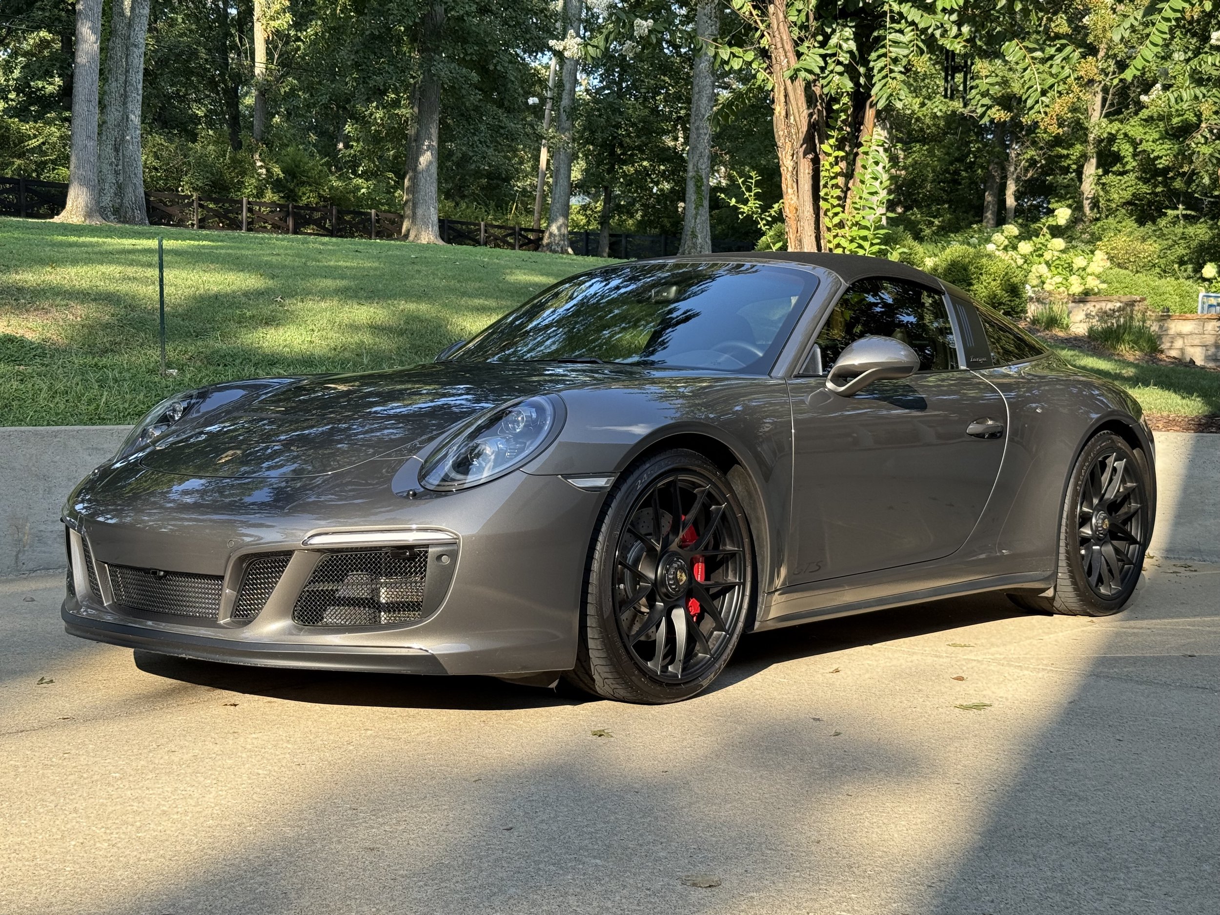 A gray Porsche 911 convertible parked on a driveway with green trees and bushes in the background.