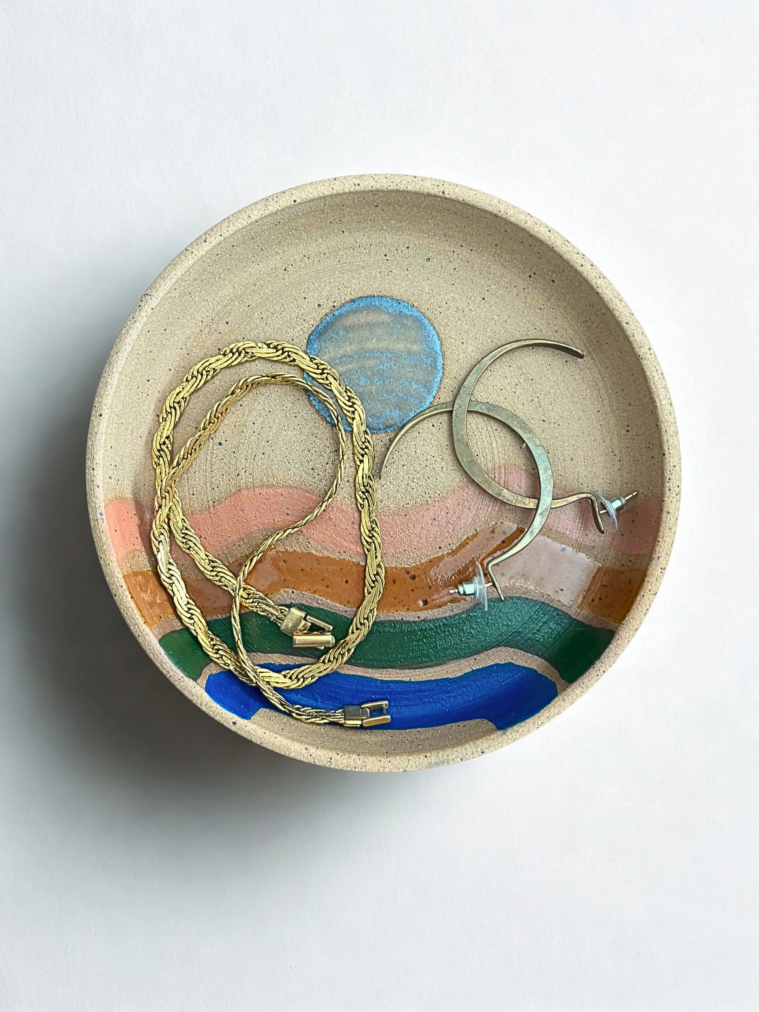 Decorative ceramic bowl with blue, pink, orange, green, and blue painted landscape, containing gold chain bracelet and silver hoop earrings.