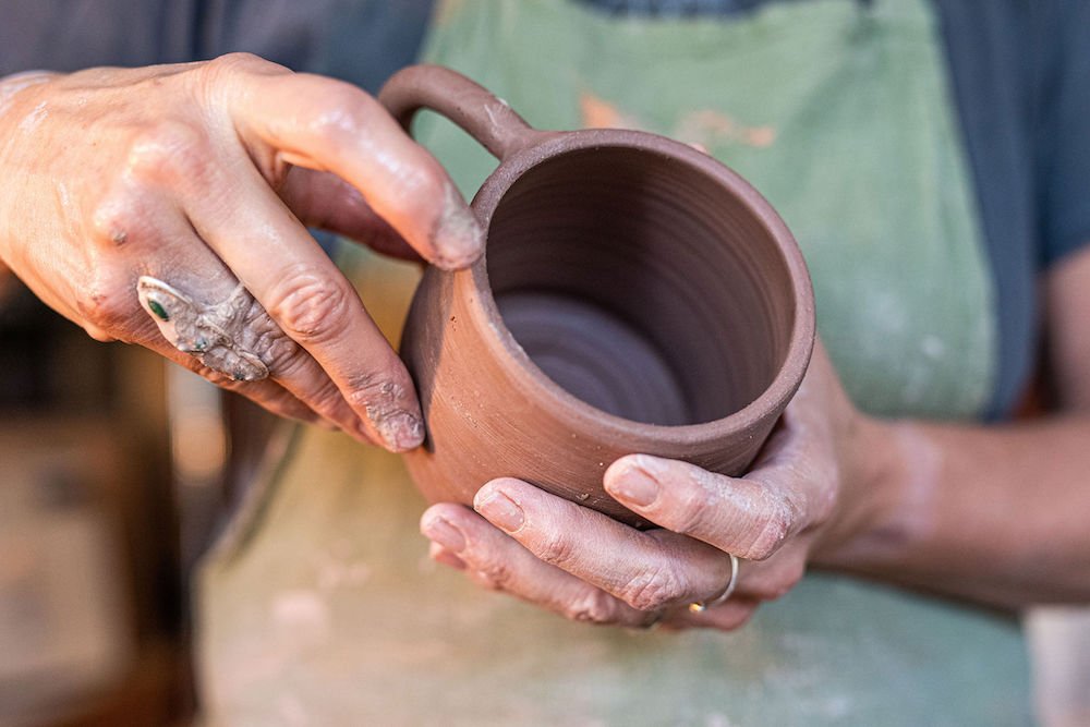 Person holding a clay mug in a pottery studio, their hands covered in clay.