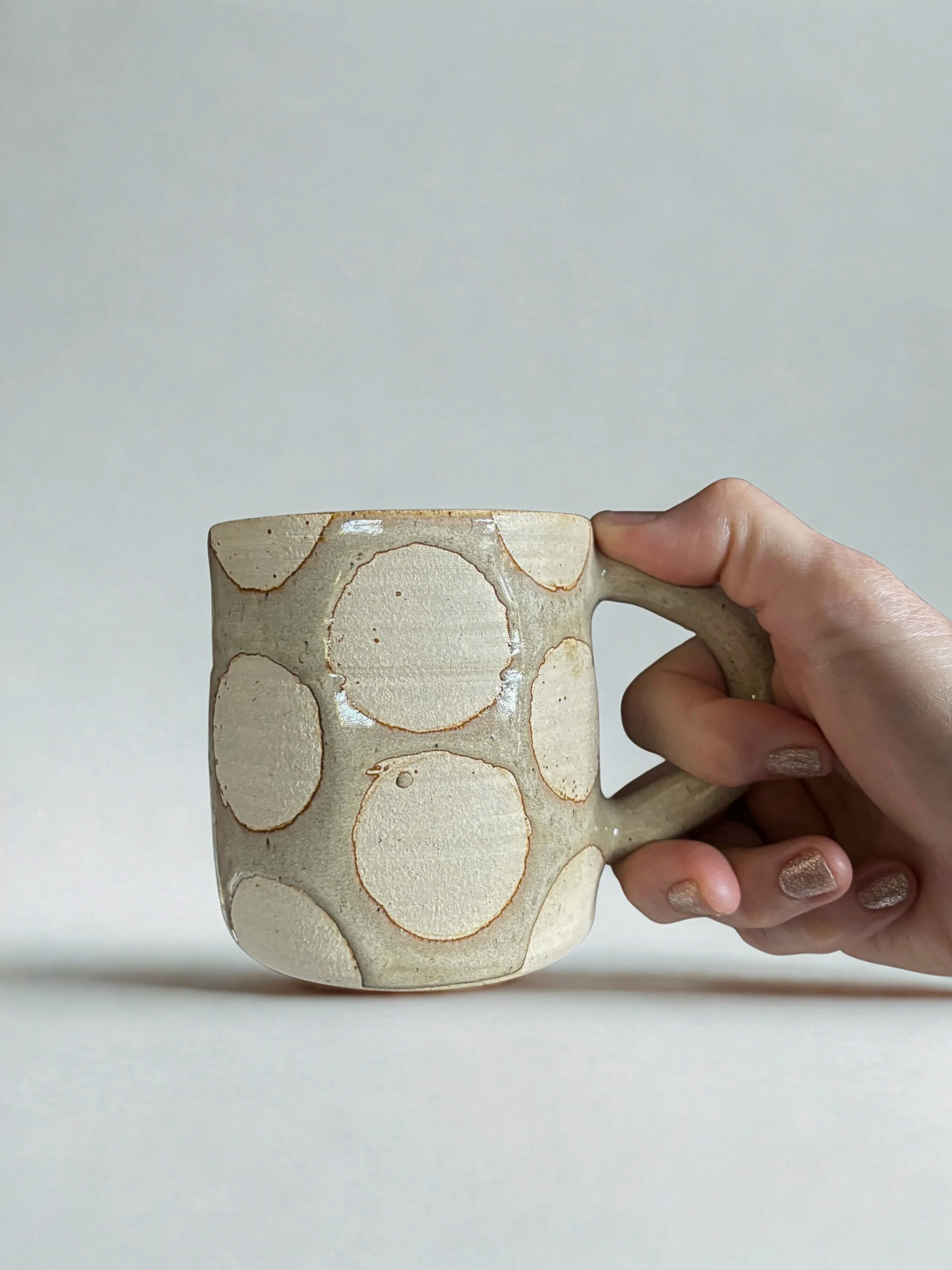 Hand holding a beige ceramic mug with a circle pattern on a neutral background.