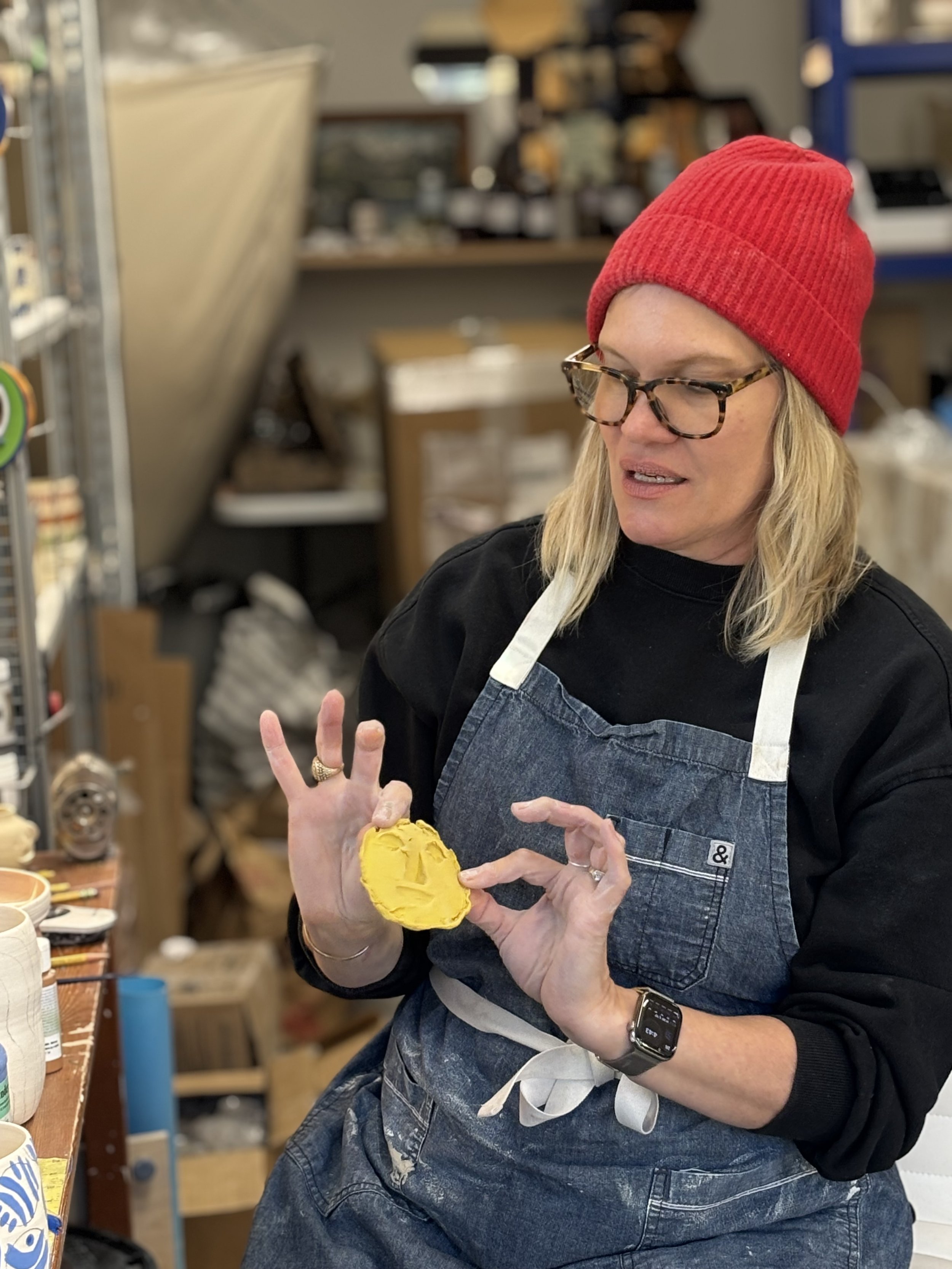 Woman with blonde hair and glasses wearing a red beanie and black shirt, holding yellow clay in a workshop.