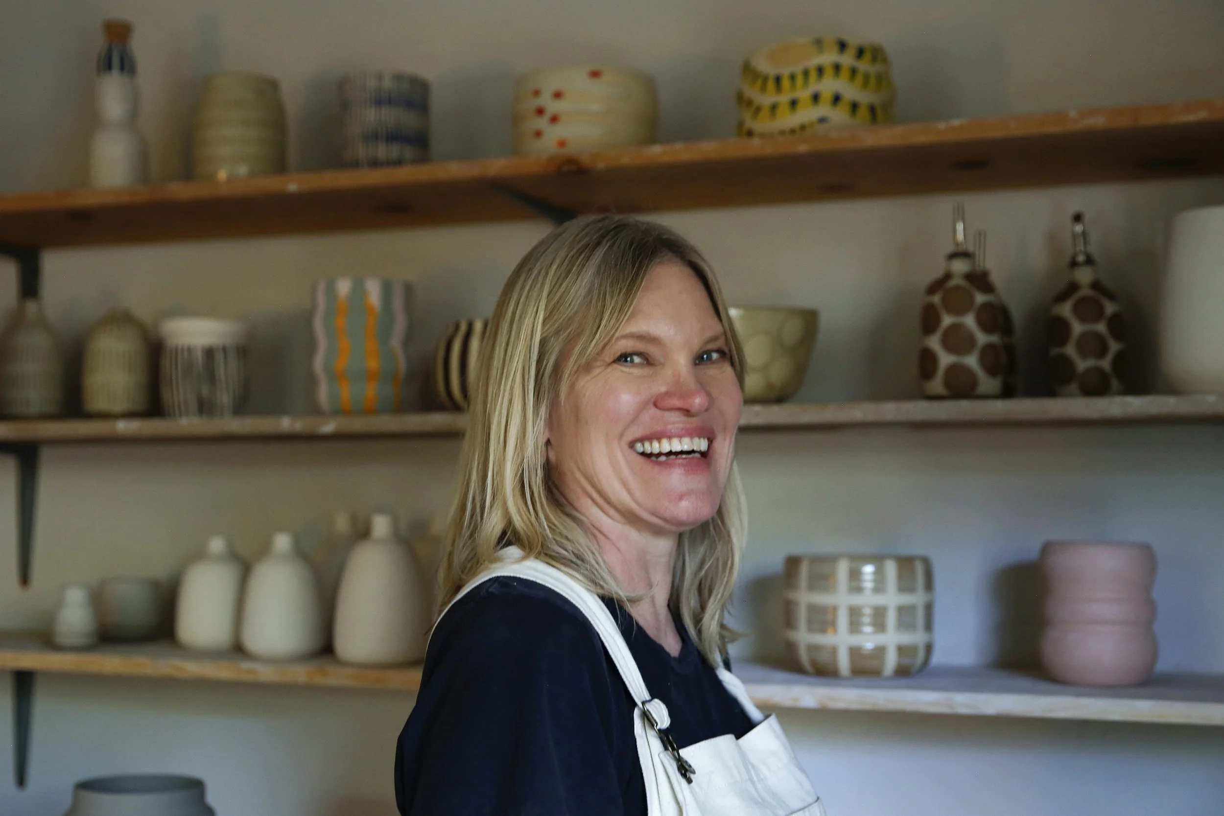 A smiling woman with blonde hair, wearing a navy shirt and an apron, standing in front of wooden shelves filled with various patterned ceramic bowls and bottles.