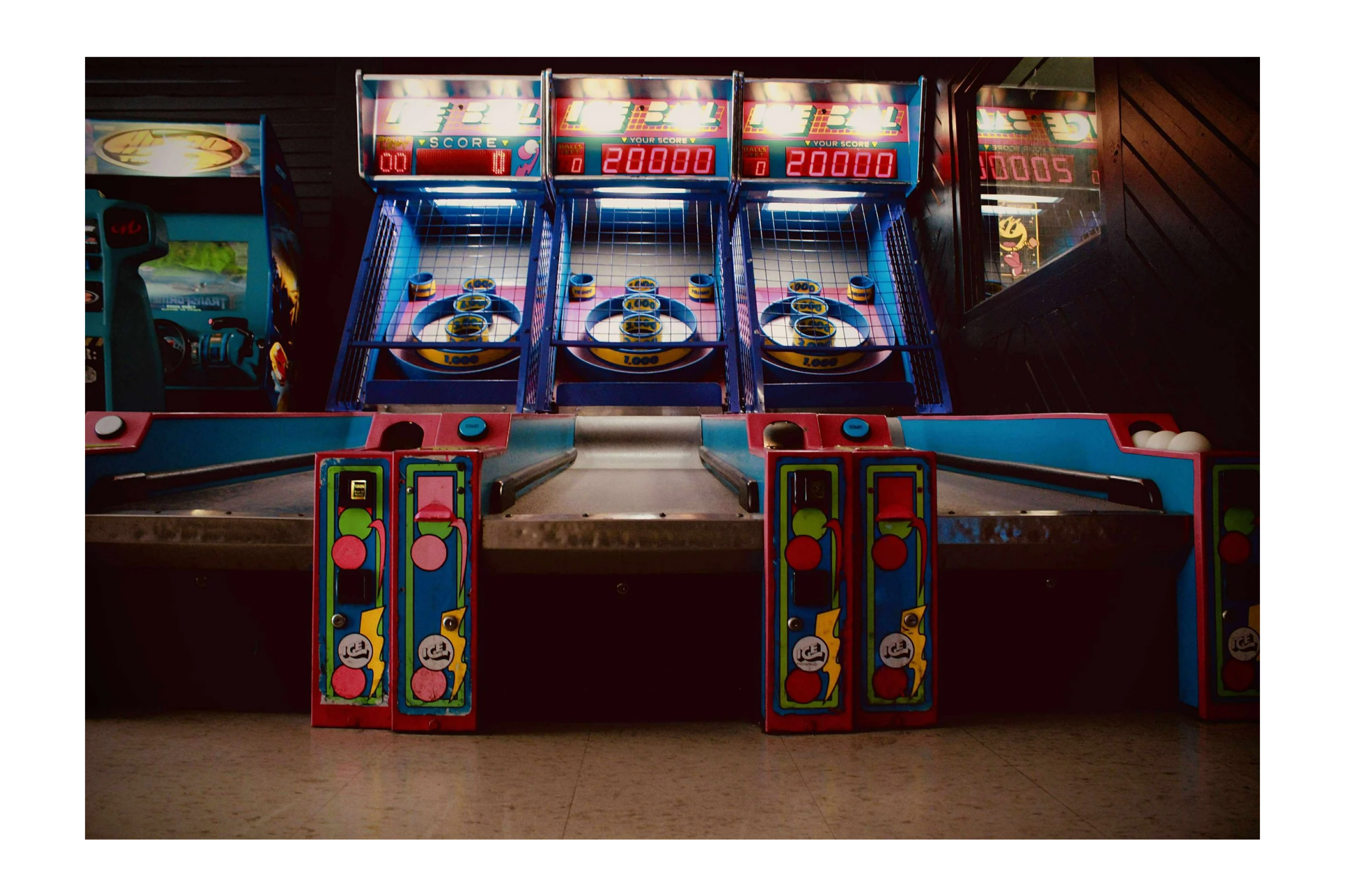 Three skee-ball machines at an arcade, with digital score displays and colorful designs, in a dimly lit room.