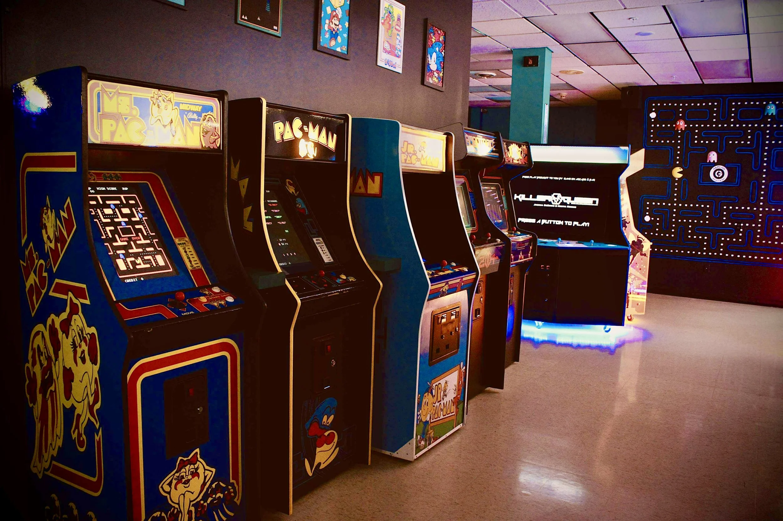 A row of vintage arcade game machines, including Pac-Man, Ms. Pac-Man, Jr. Pac-Man, and new 10 player arcade game Killer Queen. In an arcade with wall posters of popular cartoons and a Pac-Man maze wall art.