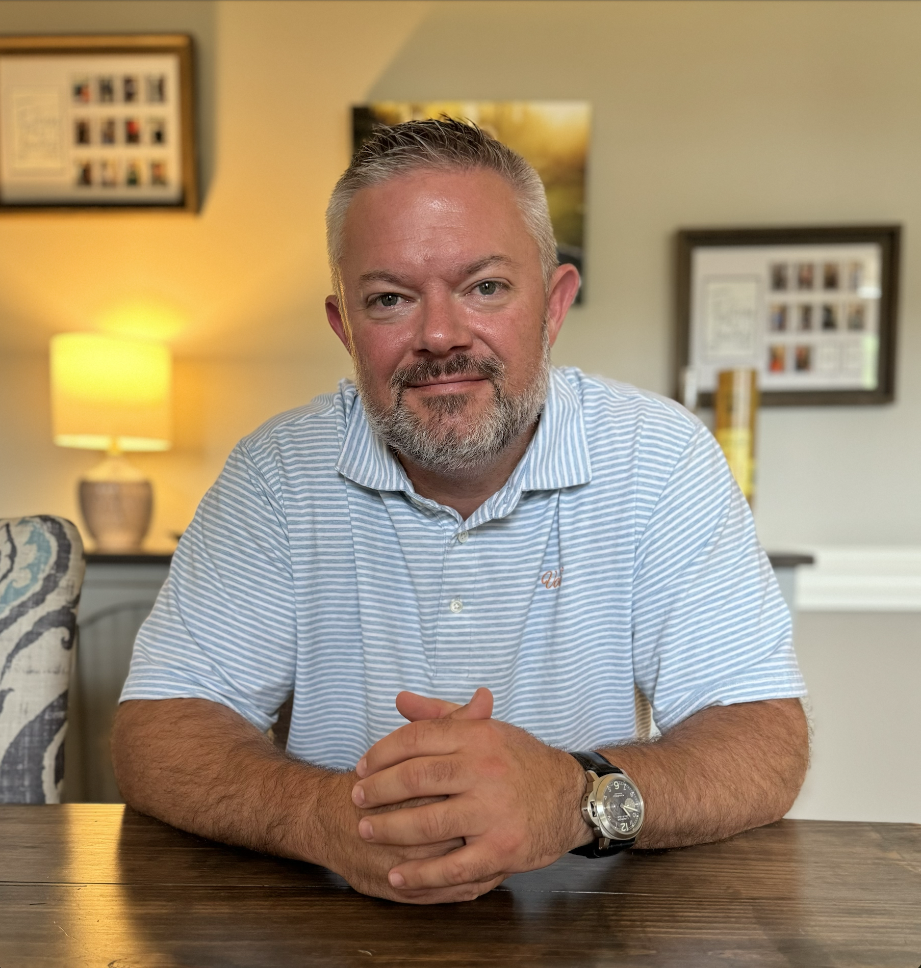 A middle-aged man with gray hair and a beard is sitting at a wooden table in a cozy living room. He is wearing a light blue and white striped polo shirt and a black wristwatch. The background shows a warm-lit lamp on a side table, framed photographs, and artwork on the walls.