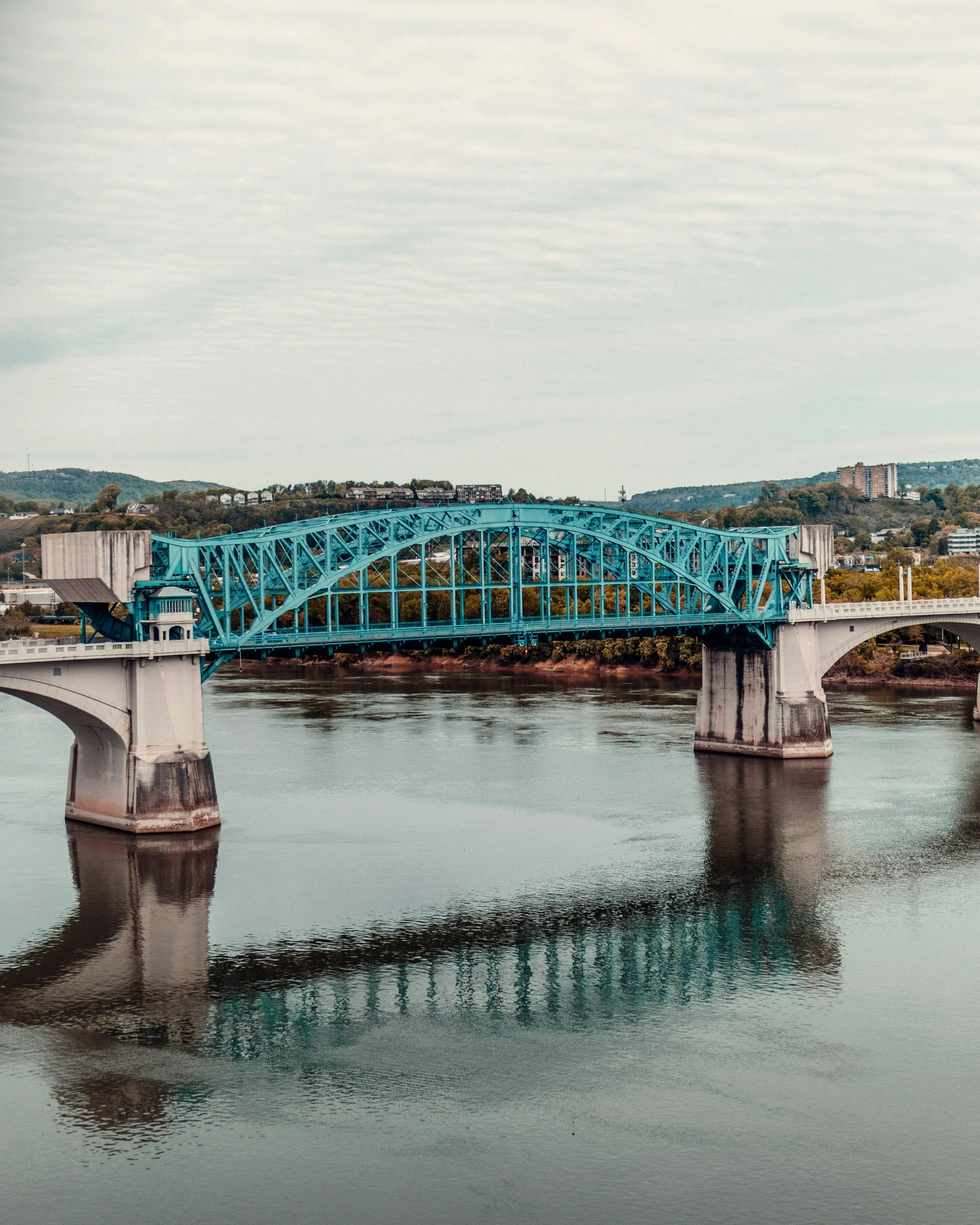 A blue truss bridge crossing a calm river with reflections, steel framework, and a distant cityscape with trees and buildings under a cloudy sky.