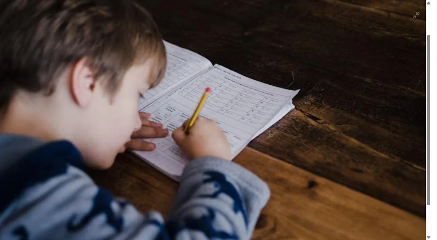 First grade boy writing math problems in a workbook at a desk