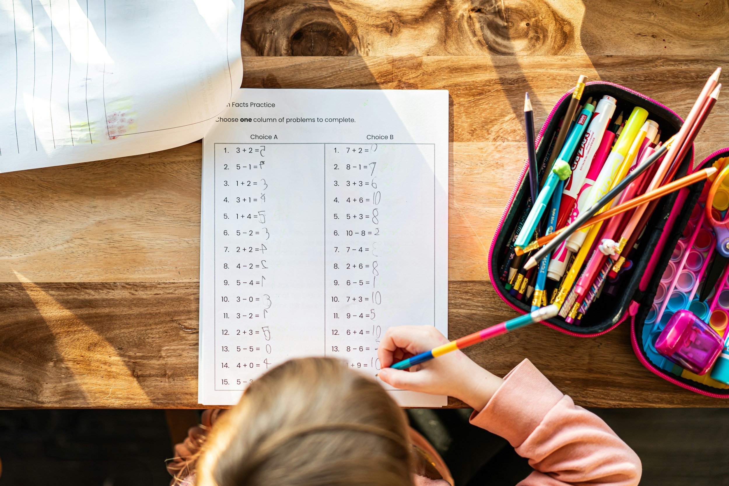 Young girl writing answers on a math worksheet at a wooden table