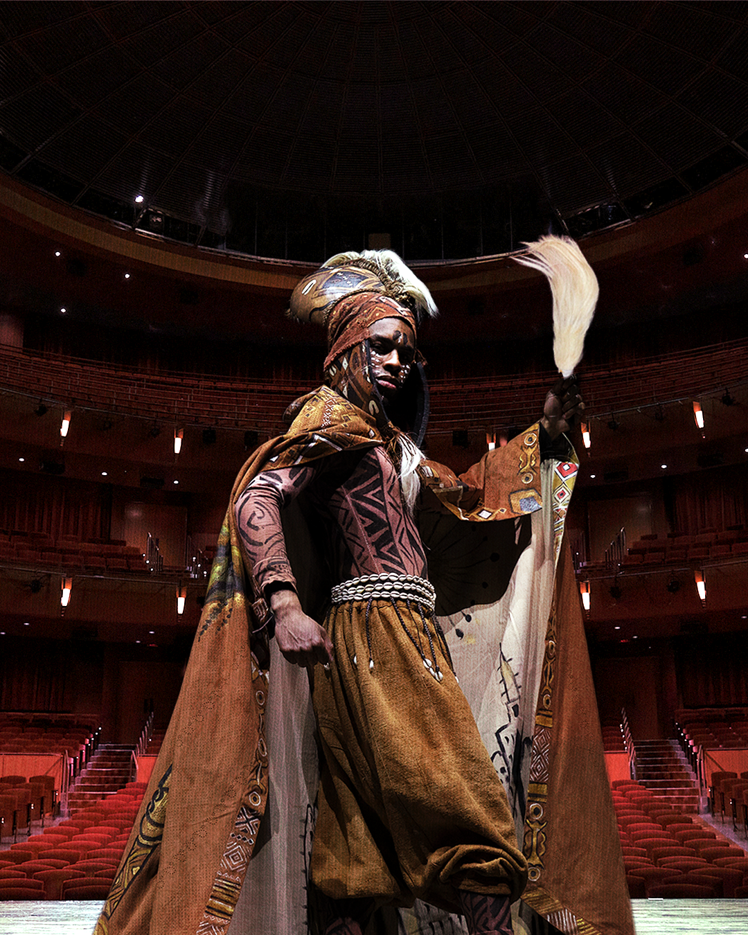 A person in elaborate traditional costume holding a feathered staff inside a large, empty theater or auditorium.