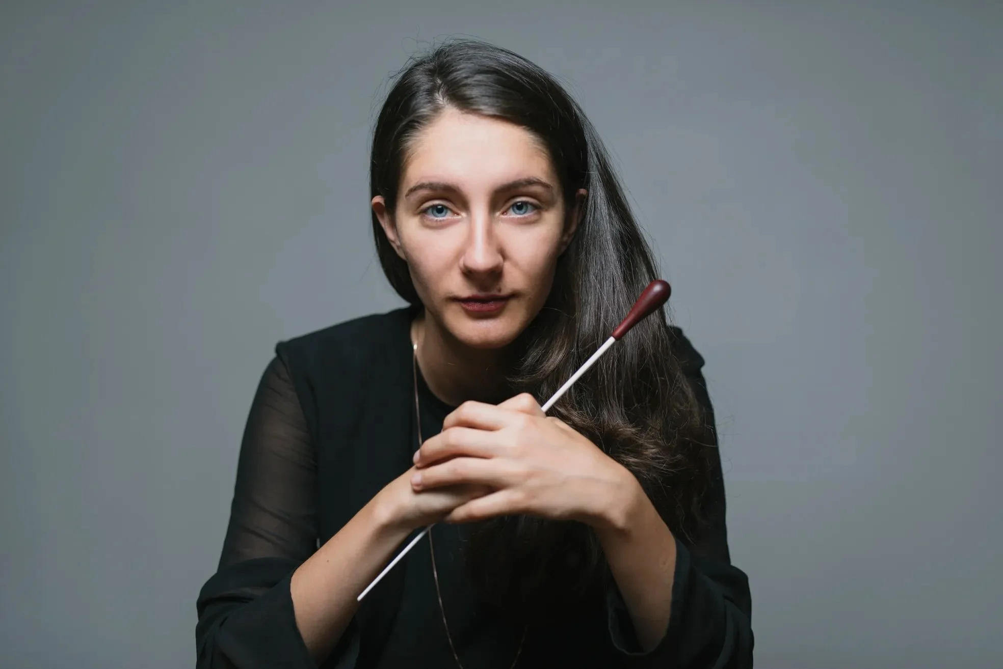 Conductor Nisan Ak, wearing a black top, holding a baton, gazing directly at the camera against a plain gray background.