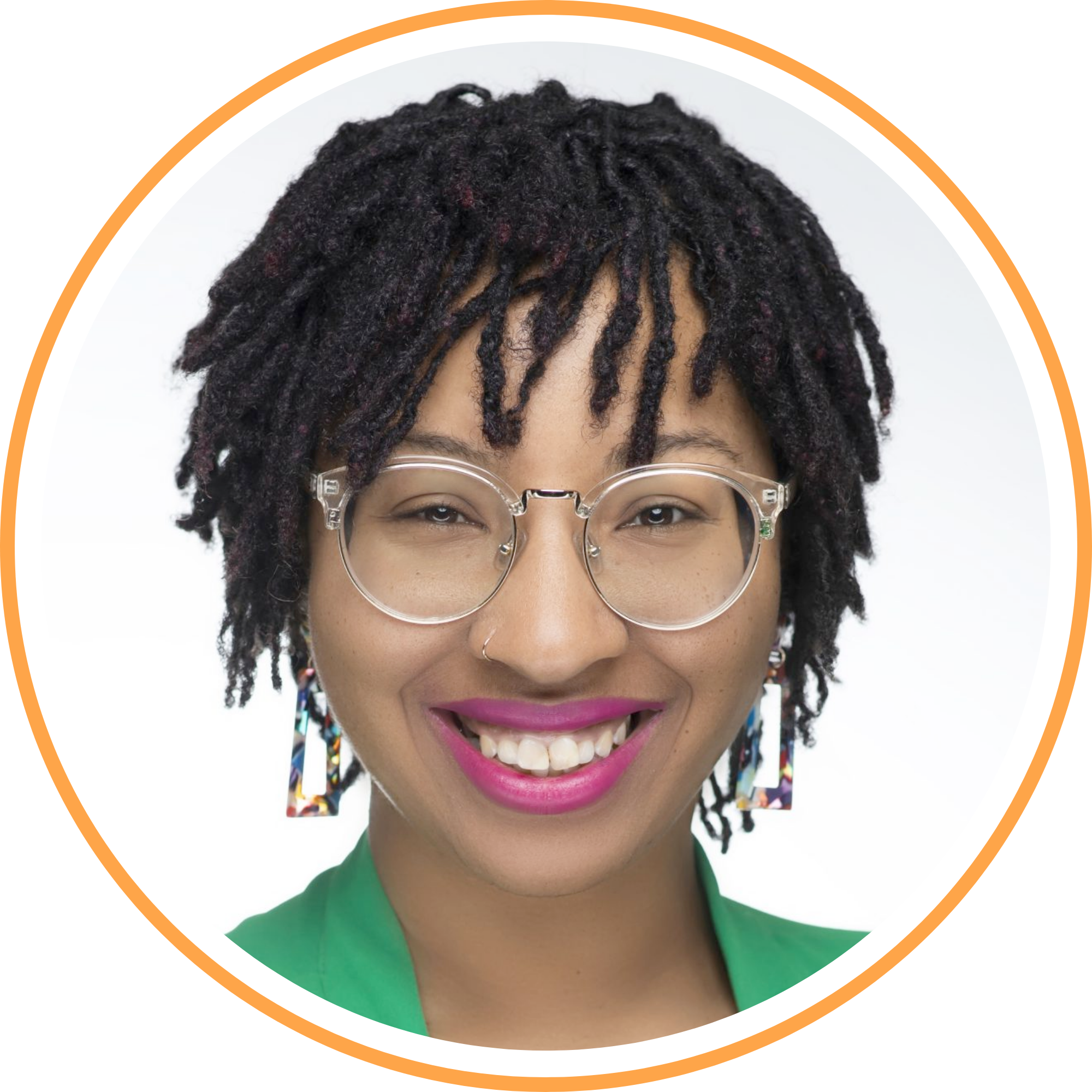 Portrait of a woman with dreadlocks wearing clear round glasses, pink lipstick, colorful earrings, and a green top, smiling against a white background.