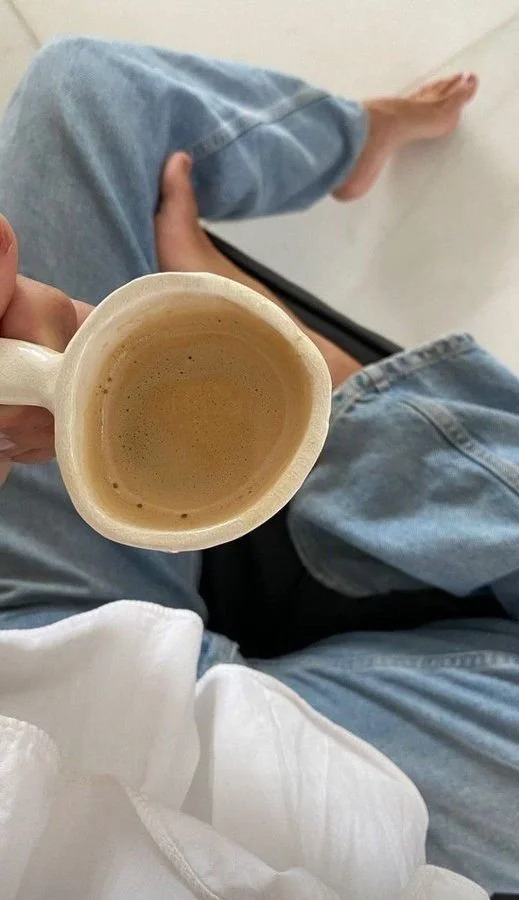 Person sitting with legs crossed, wearing blue jeans and a white shirt, holding a mug of coffee or tea, with a plain white wall in the background.