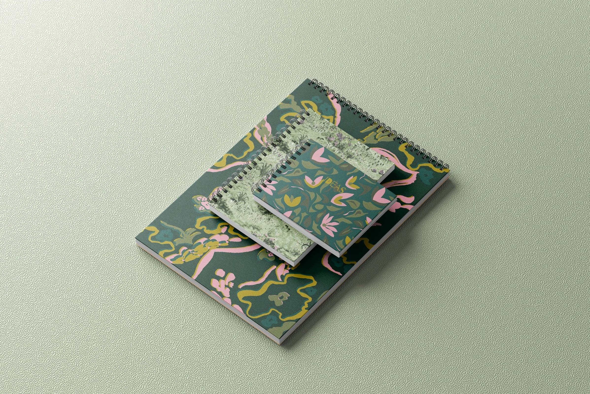 Three notebooks with floral and nature-themed covers stacked on a textured light green surface.