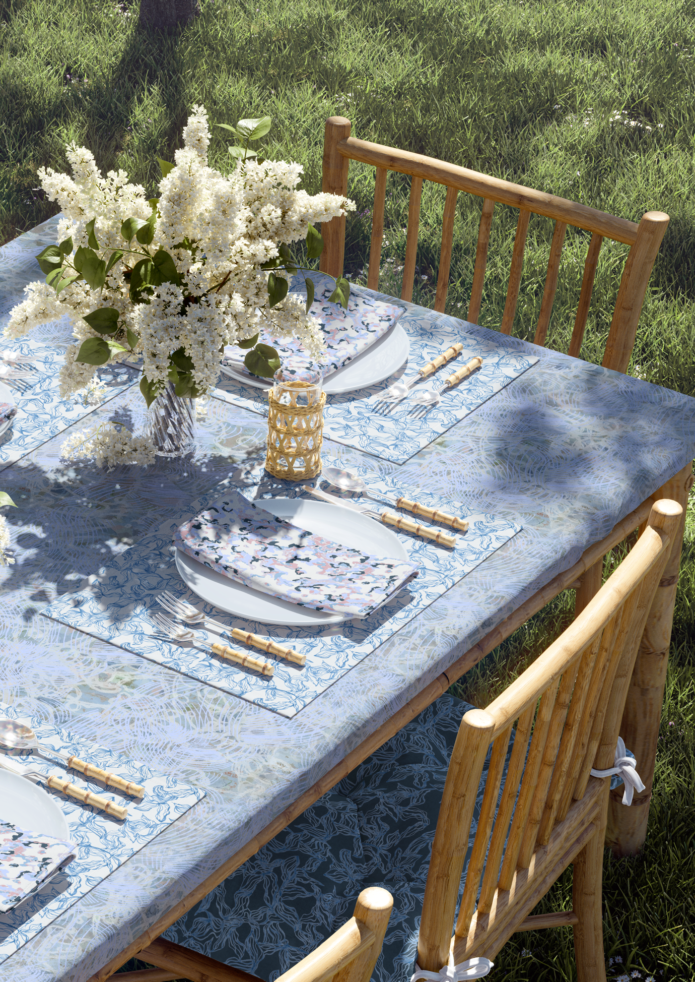 An outdoor table set for a meal with white plates, floral napkins, bamboo utensils, and a large bouquet of white flowers, on a tablecloth with a blue pattern, surrounded by wooden chairs on grass.