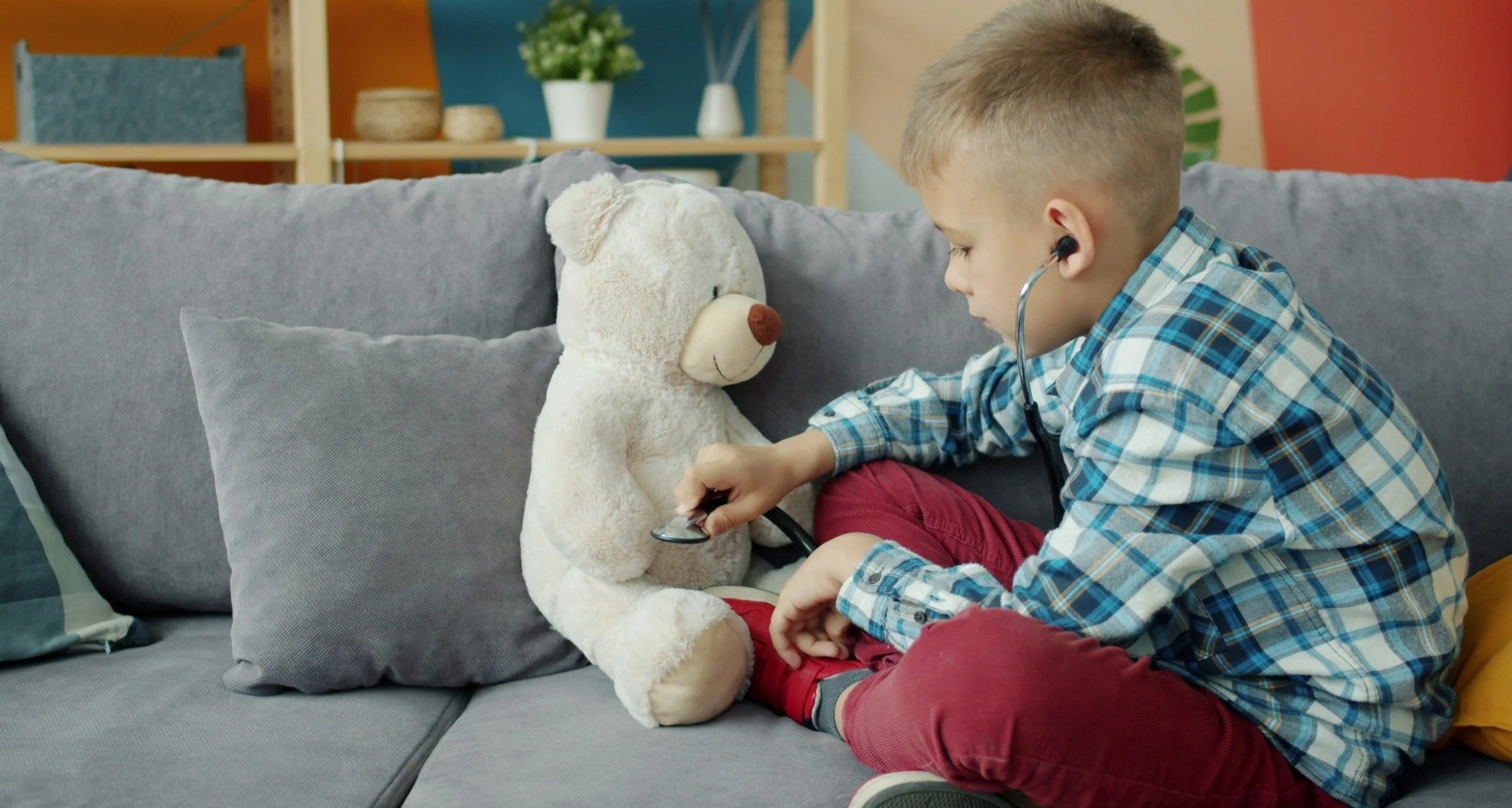 Young boy using a stethoscope on a teddy bear on a gray couch in a living room.