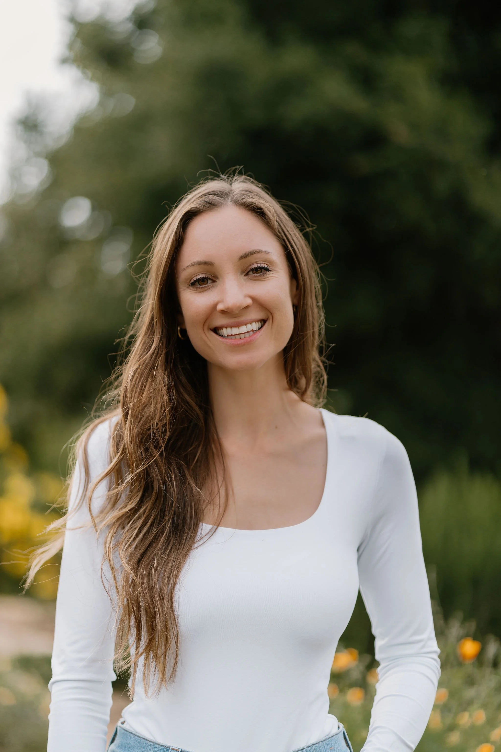 A young woman with long, wavy brown hair, smiling outdoors in a park or garden setting with green trees and yellow flowers in the background, wearing a white long-sleeve top.