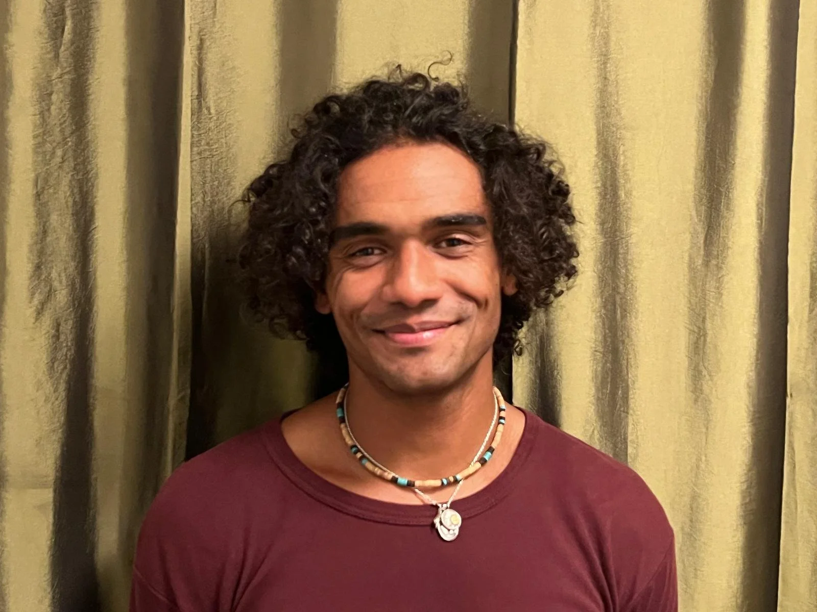 Young man with curly dark hair wearing a maroon shirt and a beaded necklace, standing in front of gold-colored curtains, smiling.