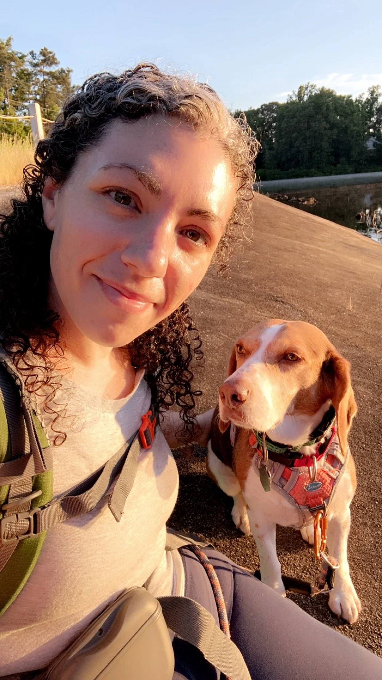 A smiling woman with curly hair taking a selfie with her dog outdoors during sunset near a body of water and trees.