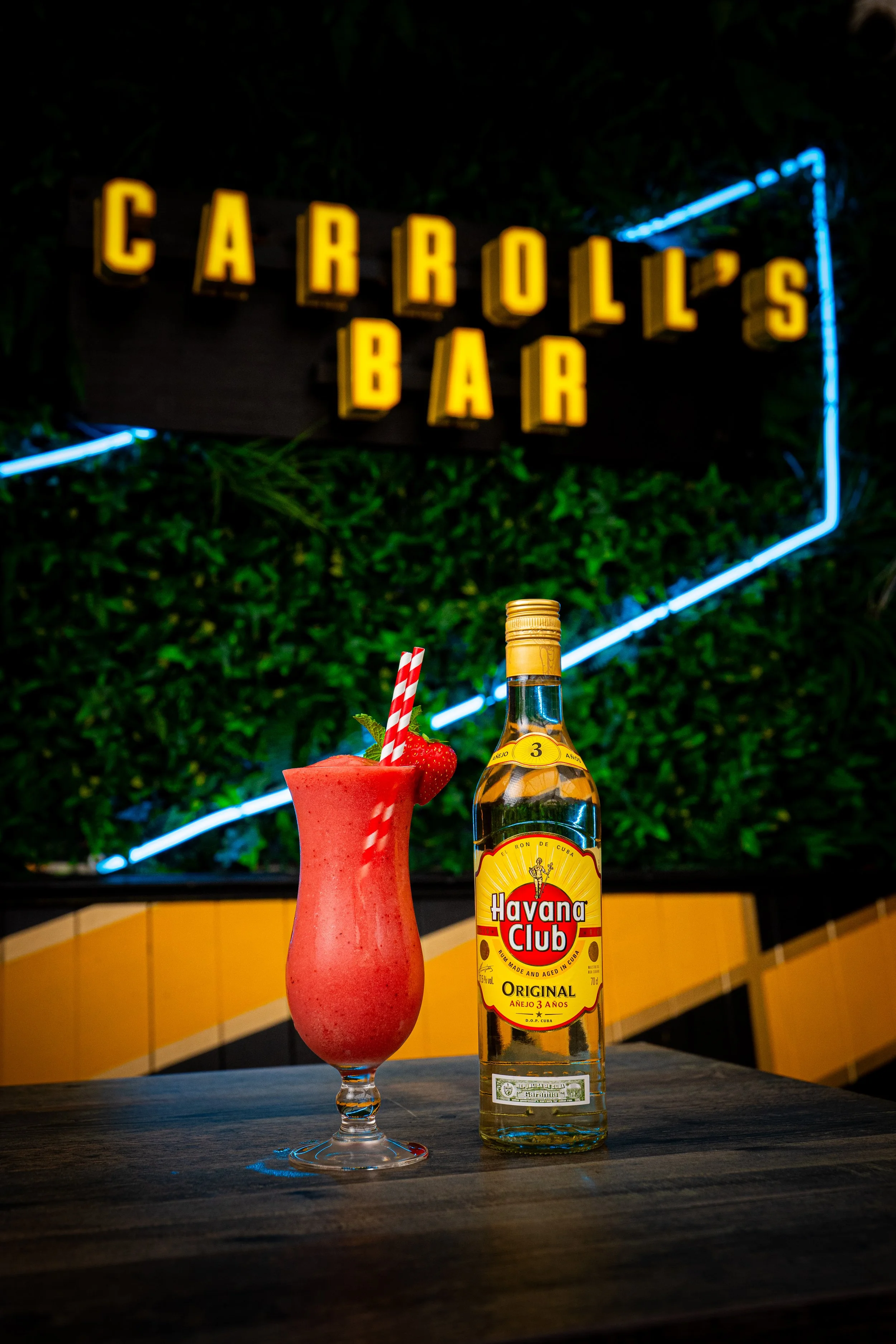 A tropical cocktail with a strawberry and striped straw next to a bottle of Havana Club rum on a wooden table. In the background, a neon sign reads 'Carroll's Bar' against a green leafy wall.