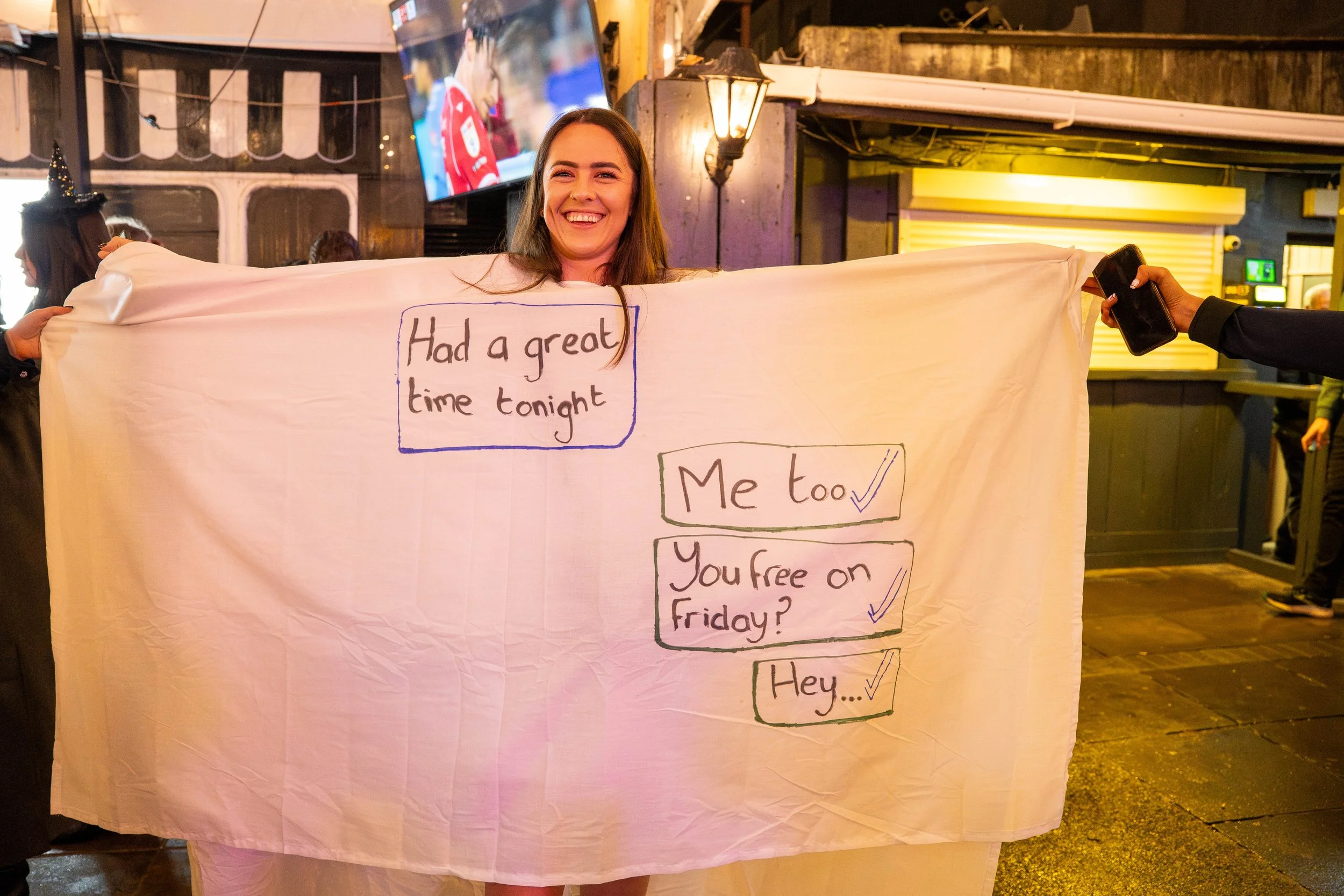 A smiling woman holding a large white blanket with handwritten messages and check marks in a lively pub or bar setting. The blanket has text that reads, "Had a great time tonight," "Me too," "You free on Friday?" and "Hey..."