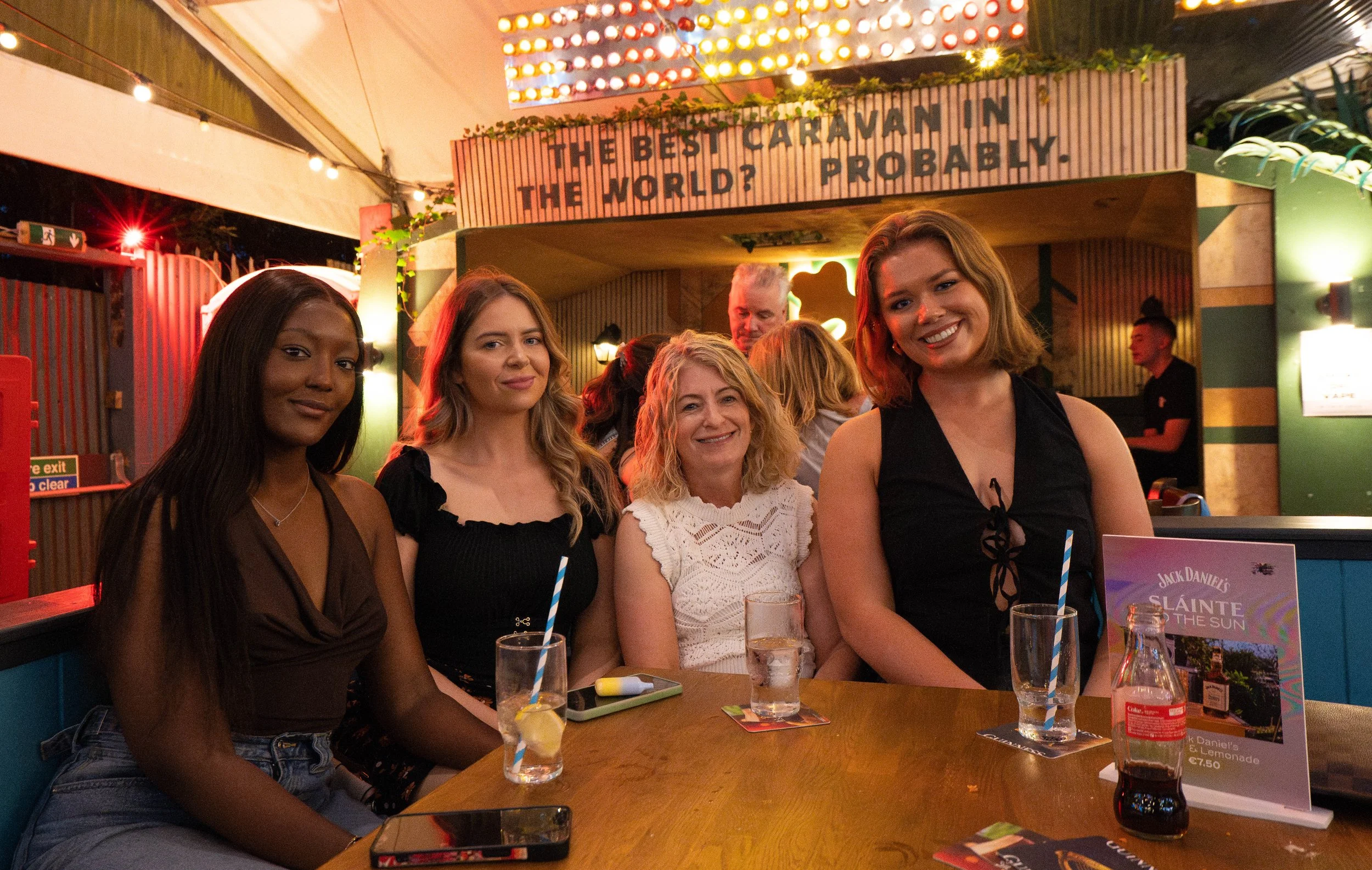 Four women sitting together at a table in a lively, colorful bar or restaurant, smiling for the photo. The background features bright lights and a sign that says 'The Best Caraman in the World? Probably.' and other patrons.