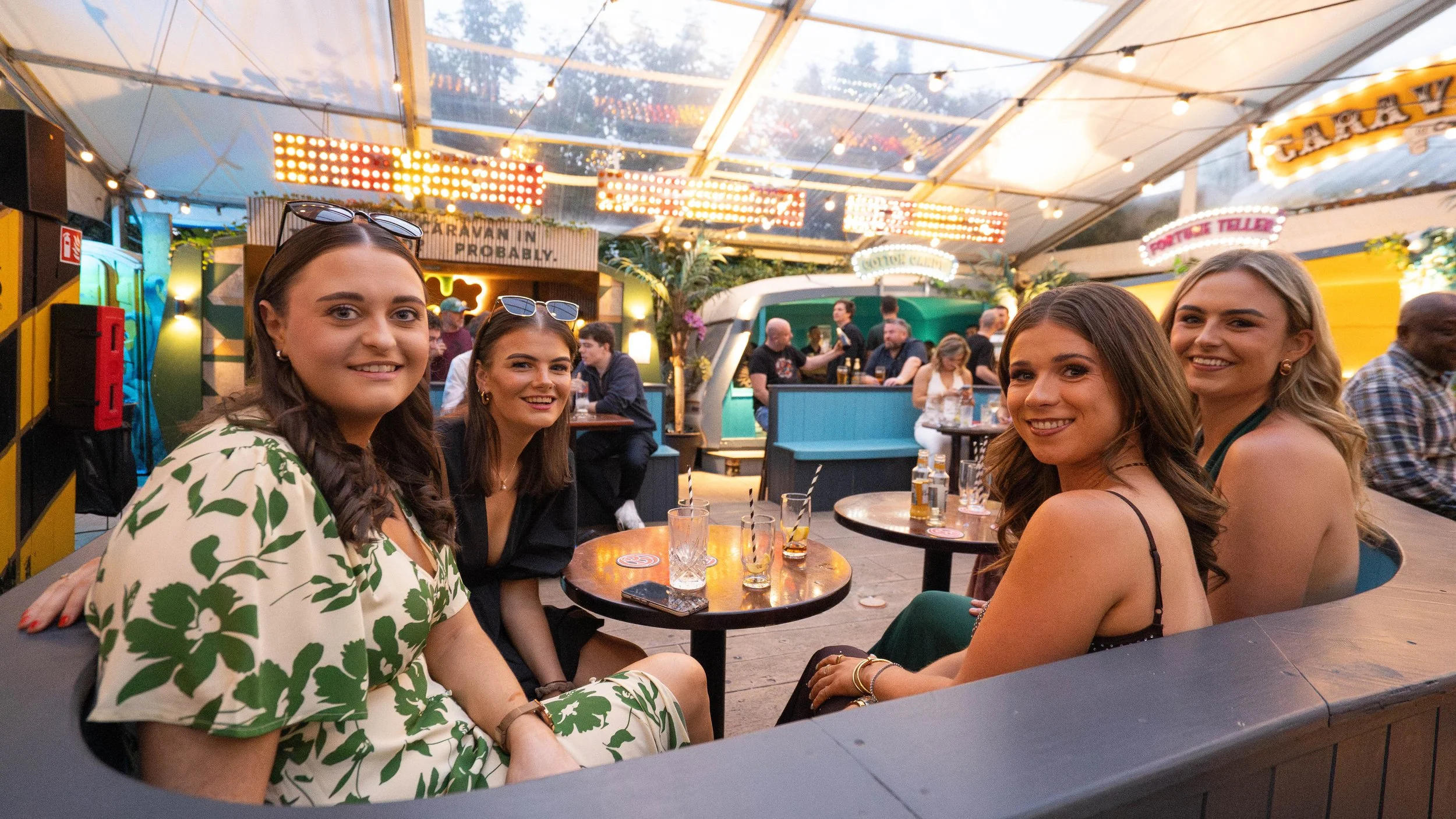 Four women smiling and sitting at a round table in a lively, brightly lit indoor venue with other people in the background.