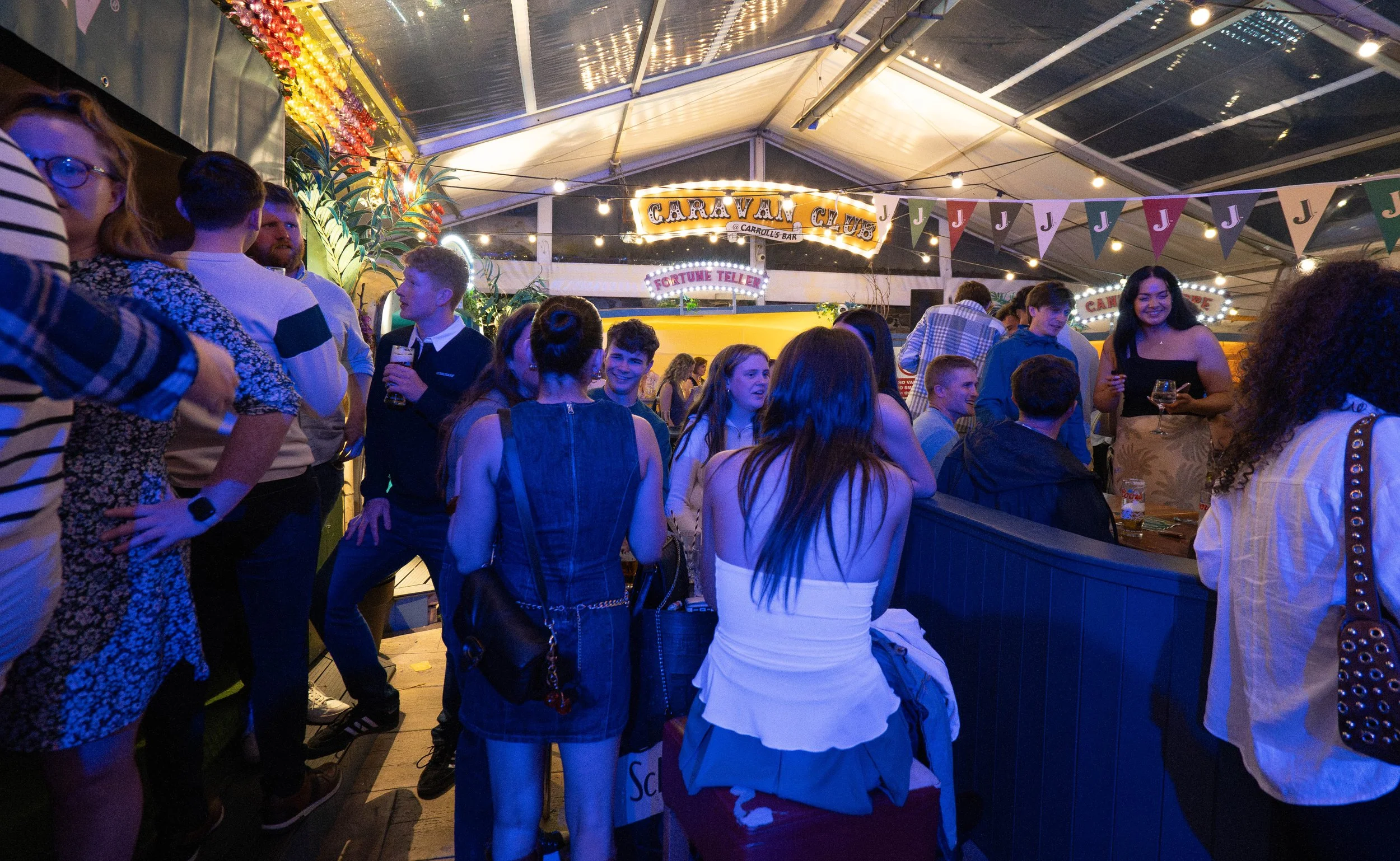 People enjoying a night at a carnival or fair inside a tent, with colorful bunting and illuminated signs for attractions like 'Caravan Games' and 'Fortune Teller' in the background.