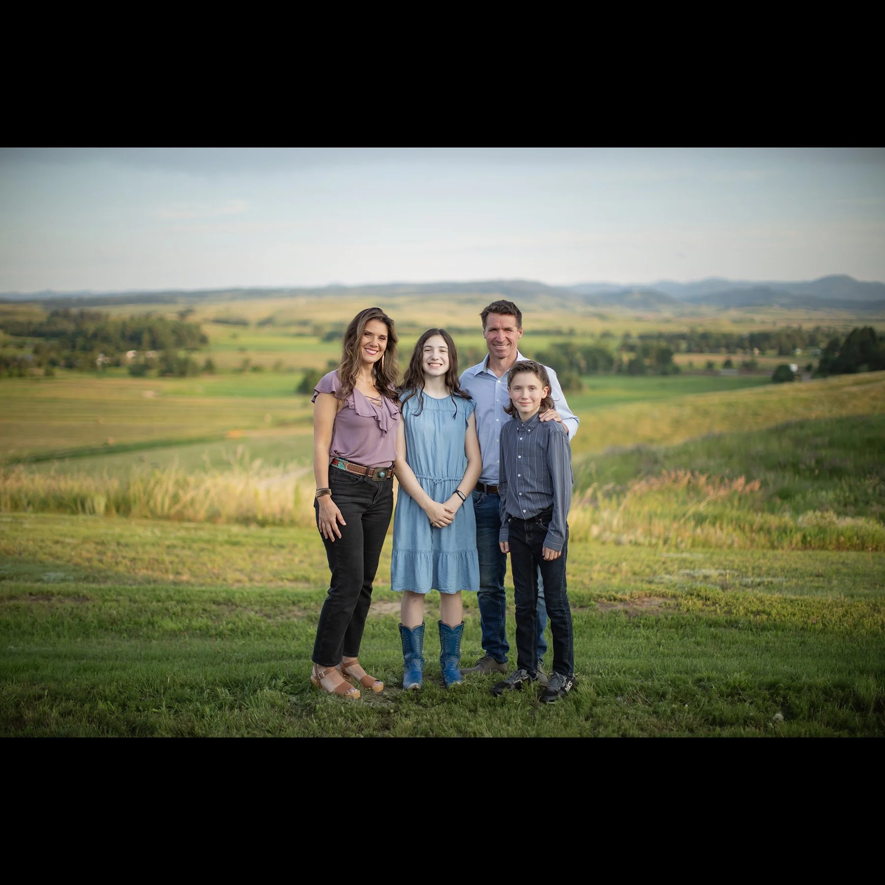 Family of four standing outdoors on grassy field with rolling hills and blue sky in background